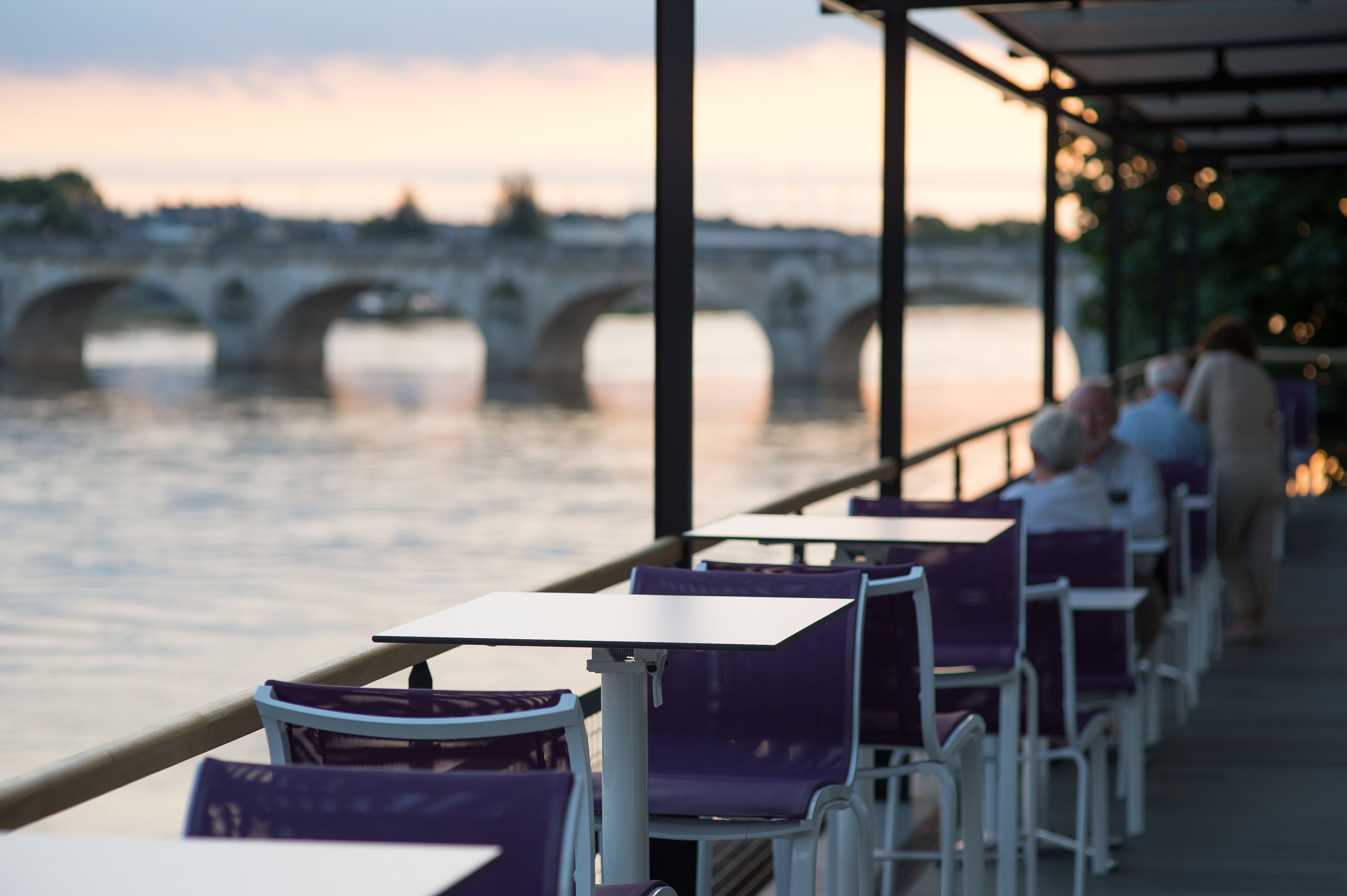 Een restaurant aan de rivier met terras, een brug op de achtergrond en uitzicht op kalm water.