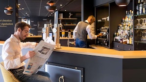 Zona de bar con mostrador, barista y un cliente leyendo un periódico.