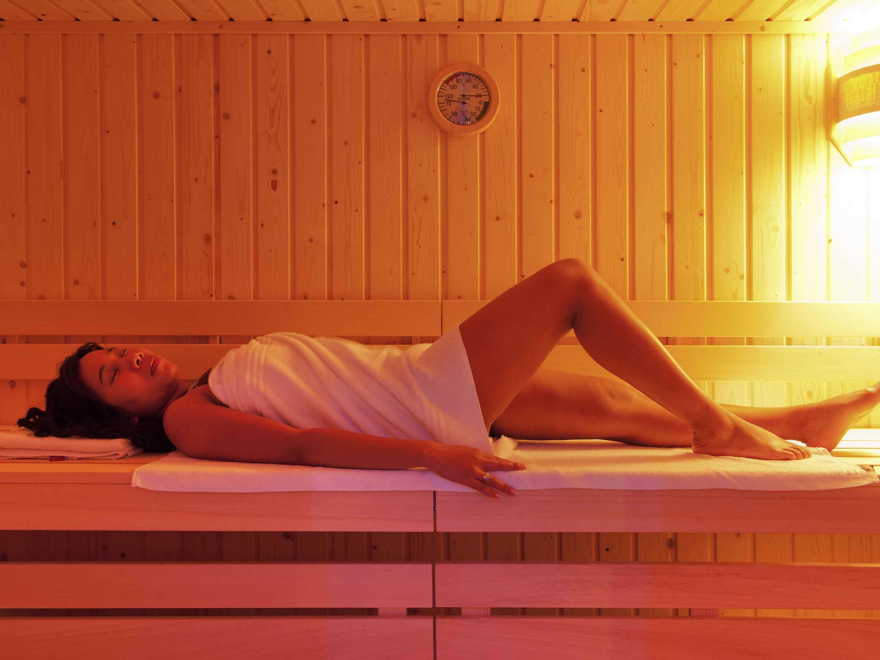 Une personne allongée sur un banc dans une pièce aux murs en bois, où l’on peut voir un thermomètre de sauna.