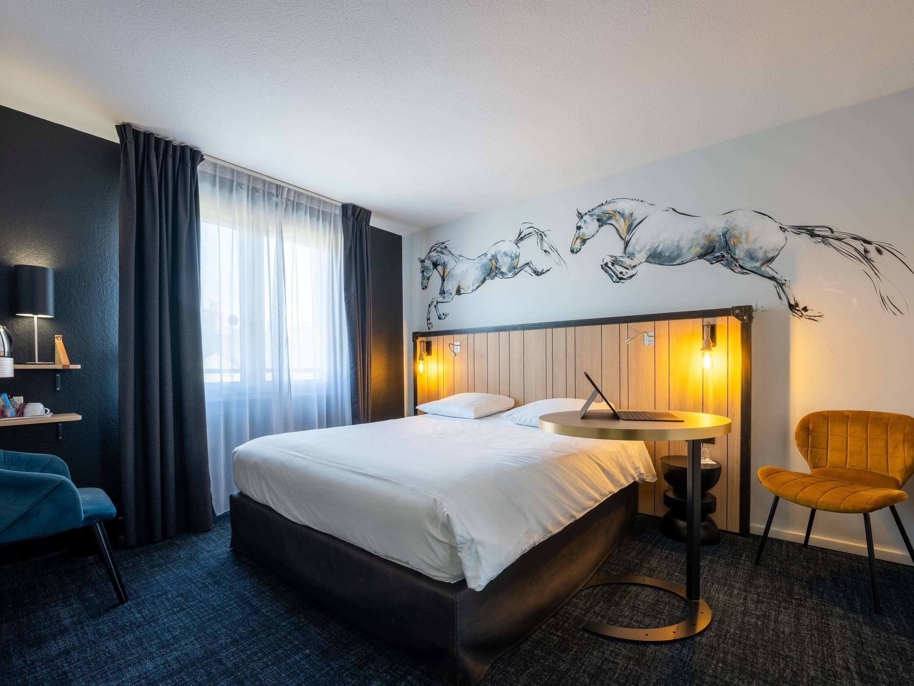 A hotel room with a bed, a desk with a laptop, a chair, a wall-mounted lamp, and a window with curtains.