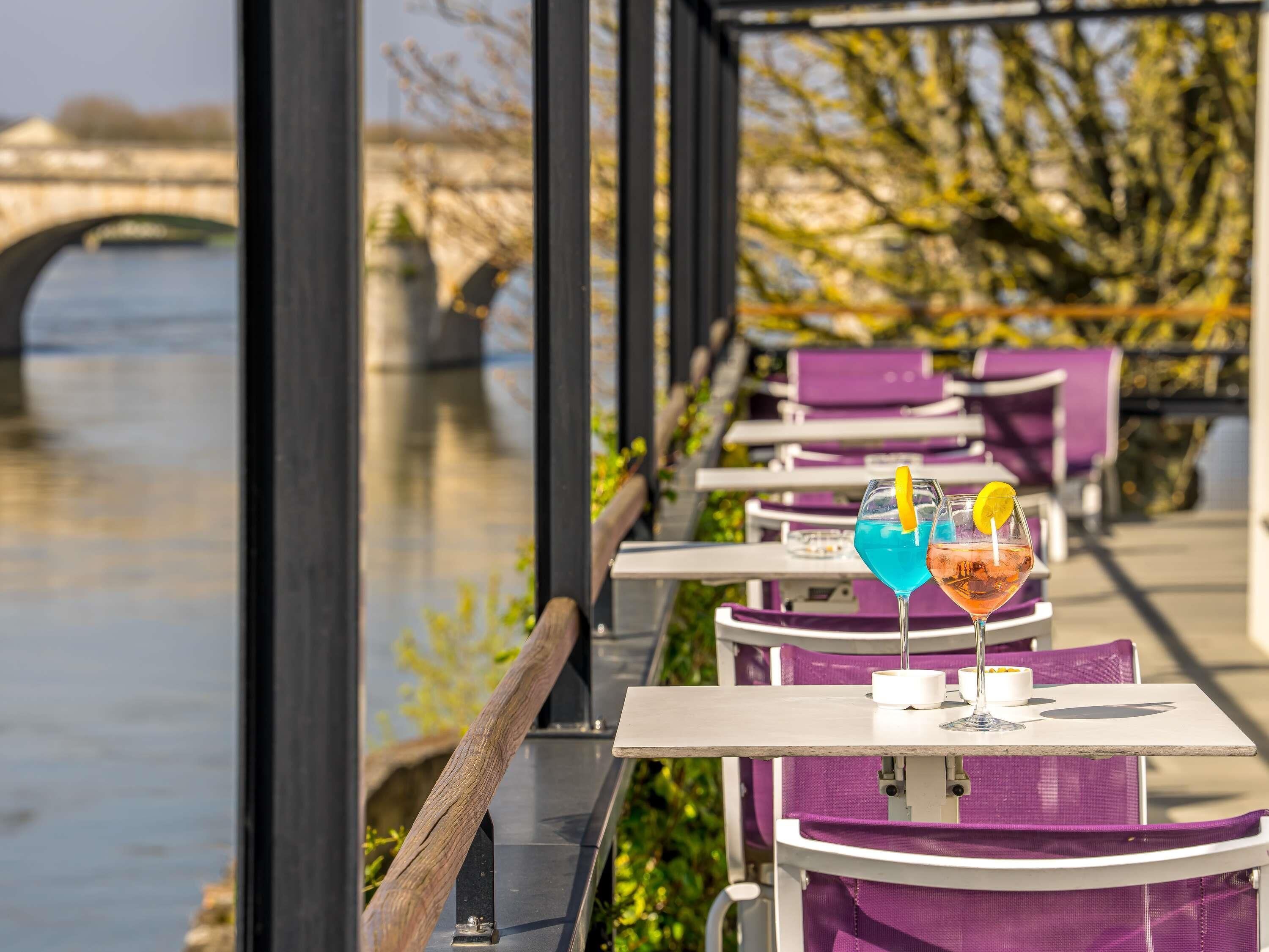 Espace extérieur aménagé avec des tables et des chaises, vue sur une rivière et un pont en arrière-plan.