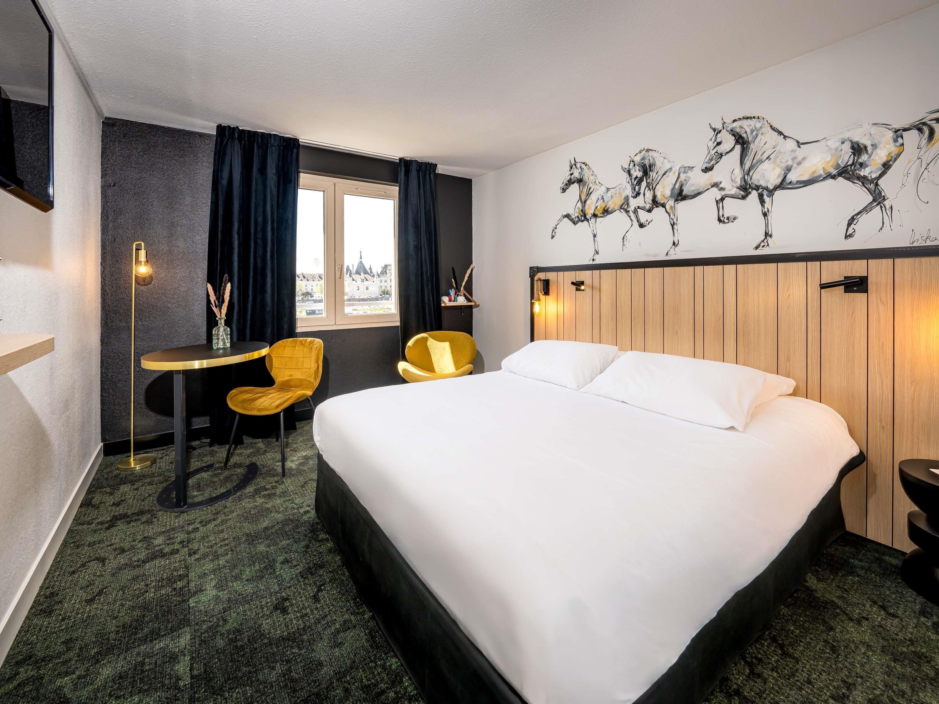 Habitación de hotel con cama, escritorio, dos sillas, ventana con cortinas y un mural en la pared con caballos.