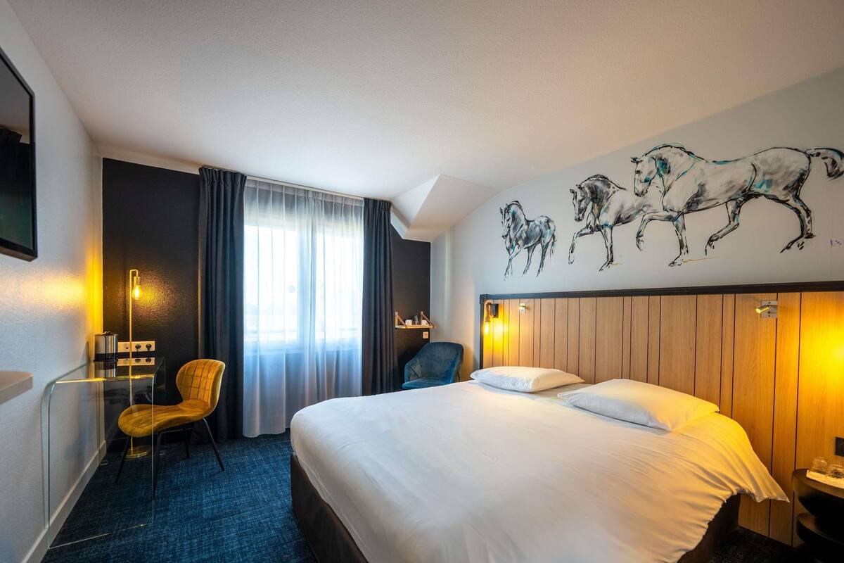 Hotelkamer met een bed, een stoel, een bureau met lamp, een raam met gordijnen en een muur met schilderijen van paarden.
