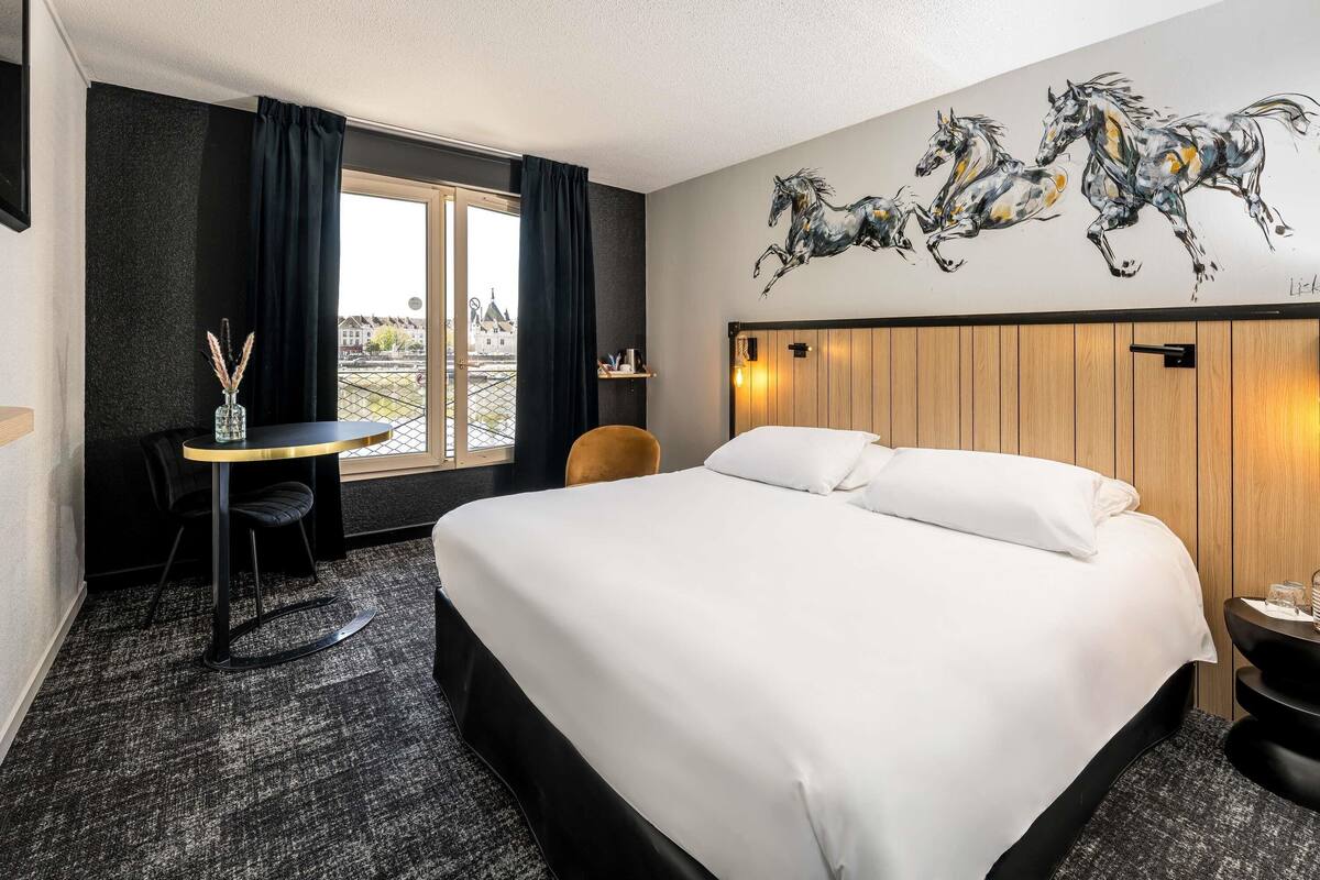 Een hotelkamer met een bed, een kleine tafel met een vaas, een stoel, een raam met gordijnen en een wanddecoratie van paarden.