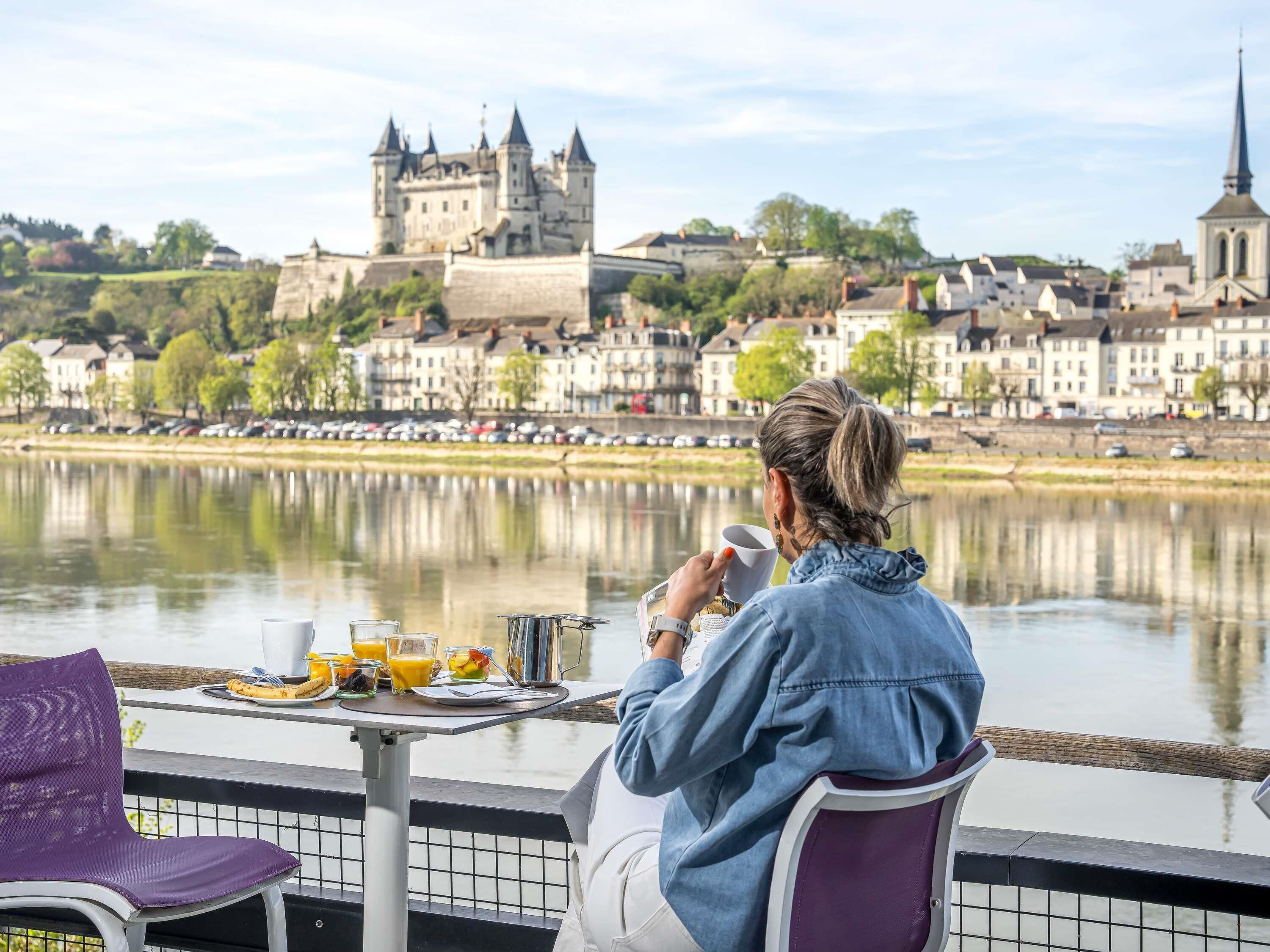 A person sitting at a table with a meal, overlooking a historic castle and river.
