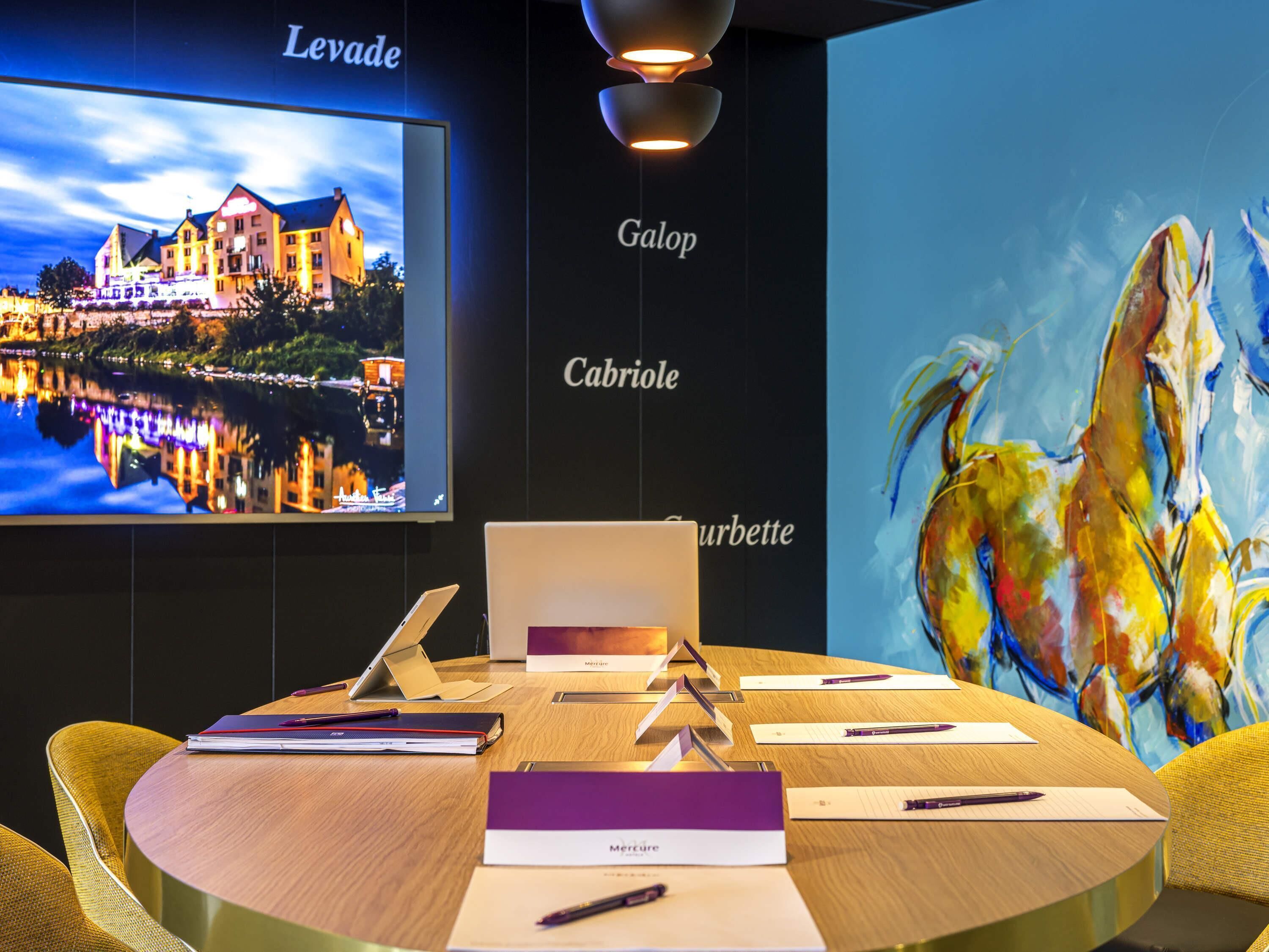 Un salón de conferencias con una computadora portátil, documentos y bolígrafos sobre una mesa, un televisor mostrando una imagen de un hotel y un cuadro abstracto y colorido en la pared.