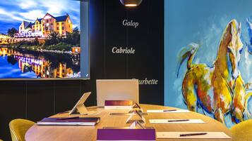 Un salón de conferencias con una computadora portátil, documentos y bolígrafos sobre una mesa, un televisor mostrando una imagen de un hotel y un cuadro abstracto y colorido en la pared.