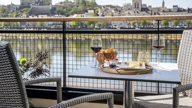Un balcón con mobiliario de mimbre, una mesa con una cesta de pan, un plato de queso y dos copas de vino tinto, con vistas a un río y un castillo con una iglesia al fondo.