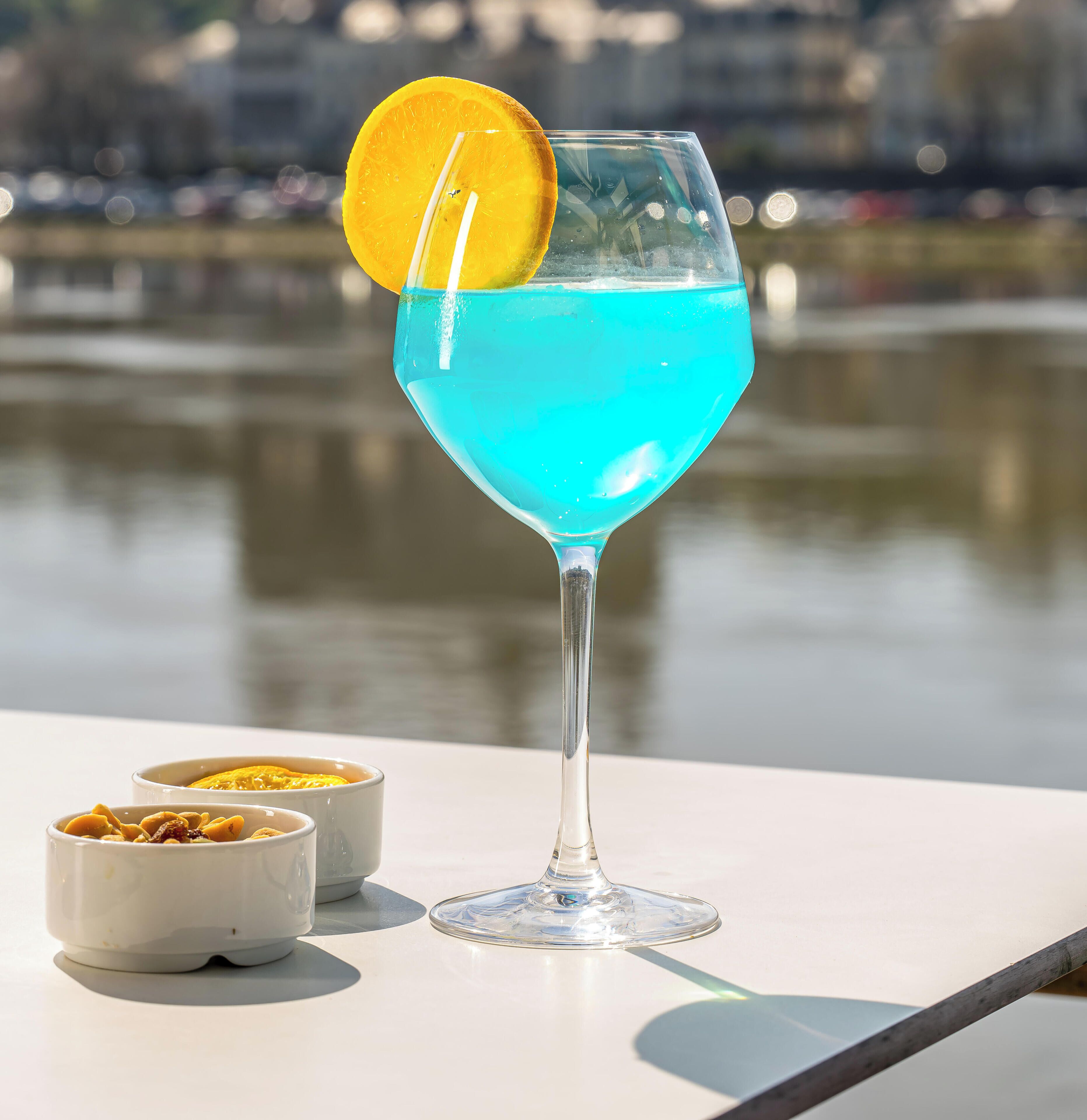 Un verre à cocktail contenant un liquide bleu et décoré d’une tranche d’orange, posé sur une table d’où l’on aperçoit un plan d’eau et des bâtiments en arrière-plan.