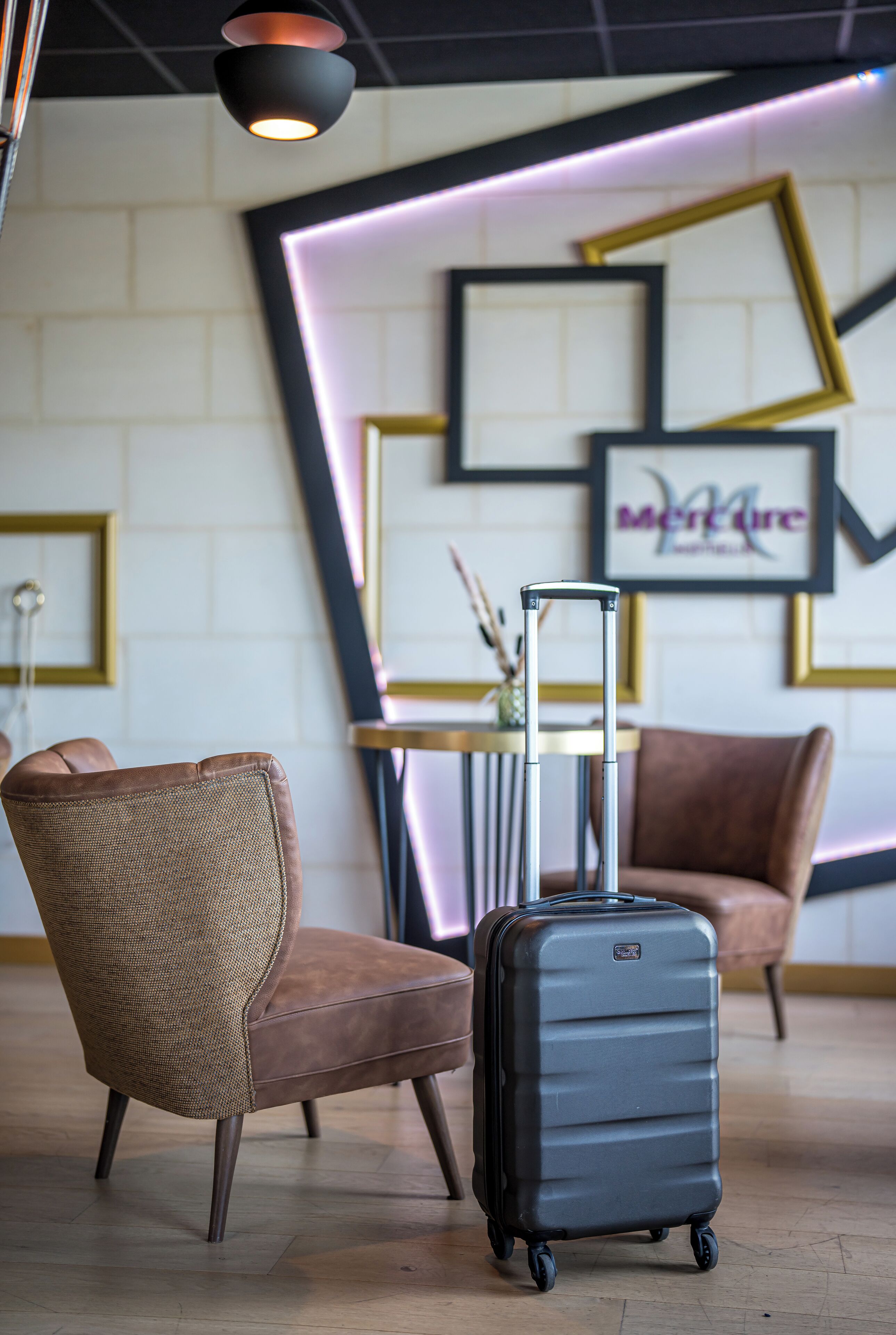 A hotel room with a brown leather chair, a black suitcase, and a table with a vase.