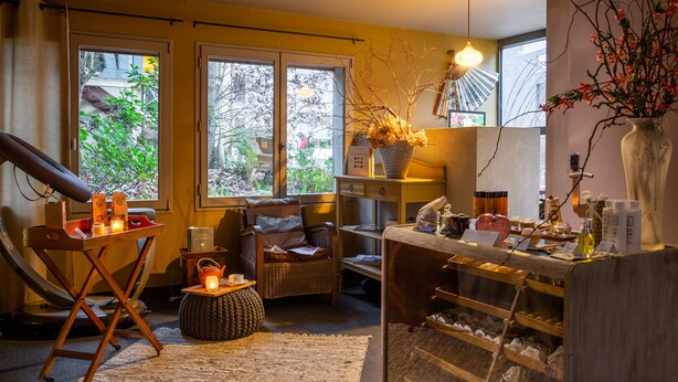 A room with a massage chair, a small table with candles, a wicker chair, a wooden counter with various items, and large windows with a view of greenery.