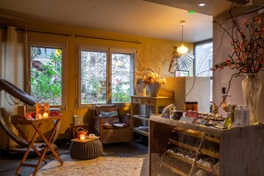A room with a massage chair, a small table with candles, a wicker chair, a wooden counter with various items, and large windows with a view of greenery.