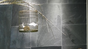 A room with dark grey tiled walls, a wicker chair with a gold cushion, and a birdcage with branches and a small bird.