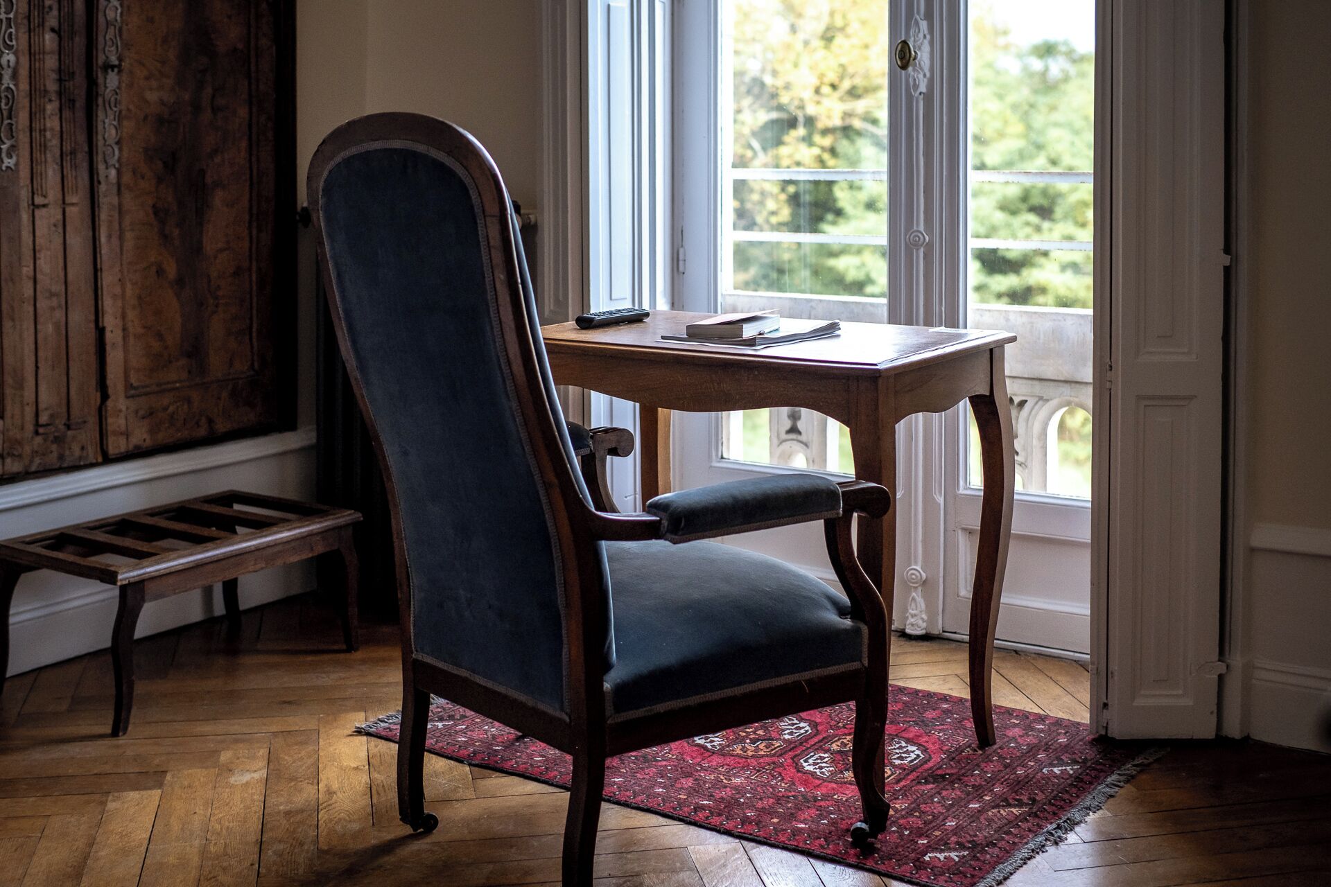 Une chaise en bois dotée d’un siège et d’un dossier rembourrés, placée devant une table en bois sur laquelle se trouvent un livre et une télécommande. La pièce est caractérisée par un plancher en bois, un tapis à motifs rouges et une vue par la fenêtre.