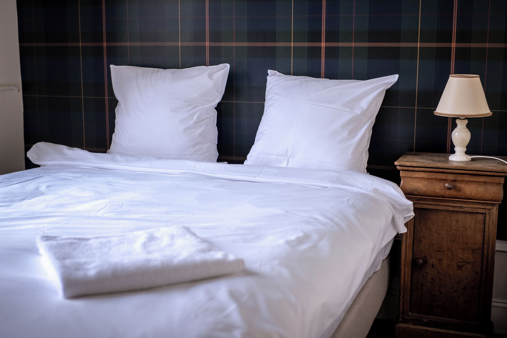 A neatly made bed with white linens and pillows, a wooden nightstand with a lamp, and a dark plaid headboard.