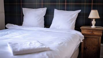 A neatly made bed with white linens and pillows, a wooden nightstand with a lamp, and a dark plaid headboard.