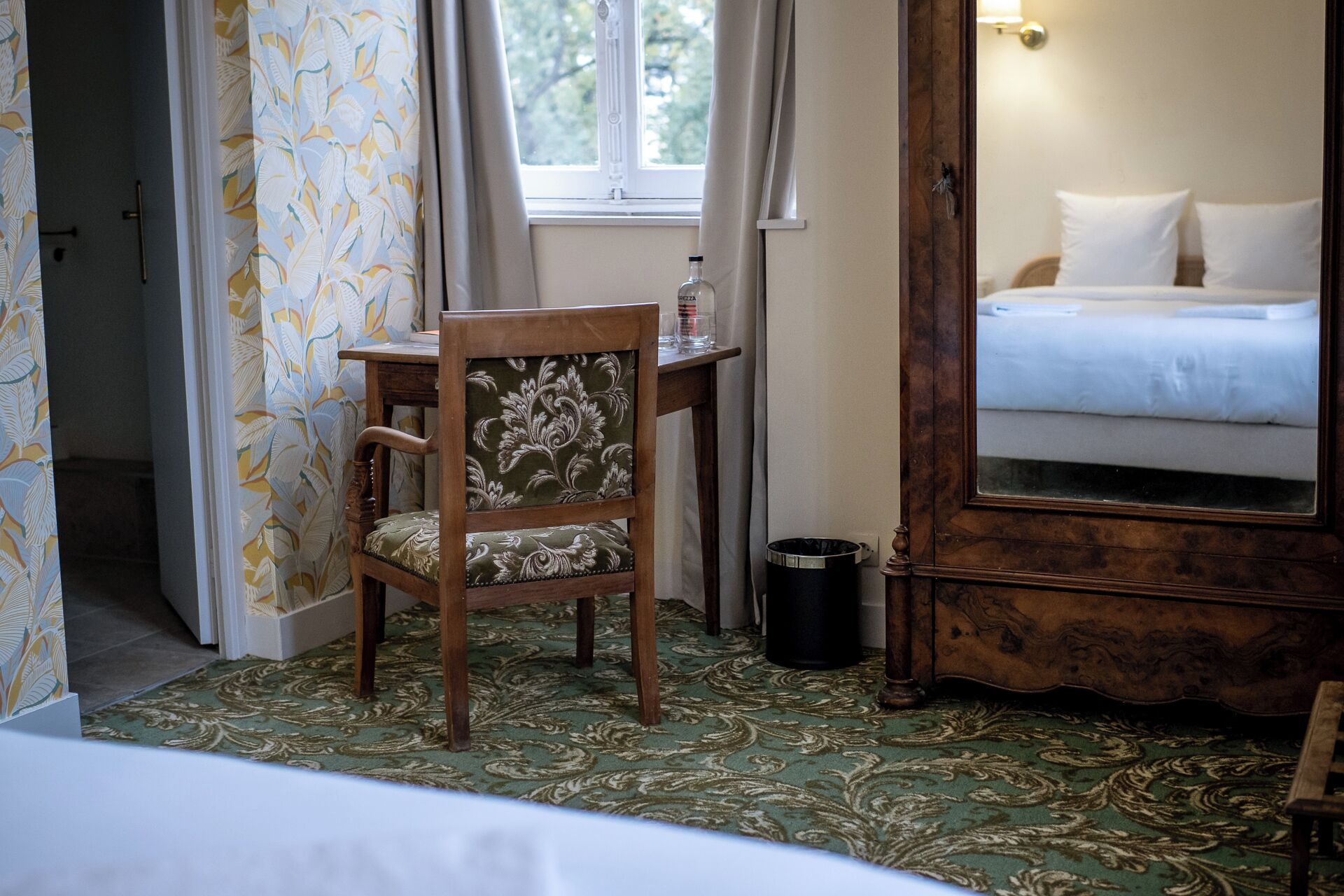 A hotel room with a bed, a wooden chair, a small table, a trash bin, and a mirror.