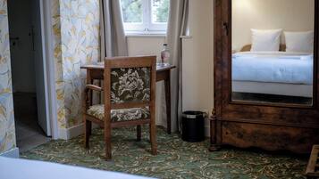 A hotel room with a bed, a wooden chair, a small table, a trash bin, and a mirror.