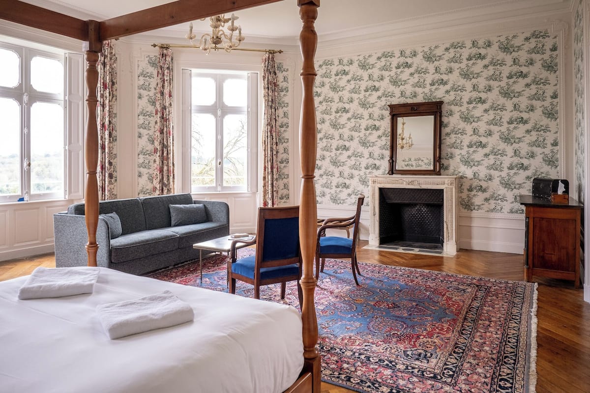 Een hemelbed met witte lakens, een grijze bank, een blauwe fauteuil en een getextureerd vloerkleed in een kamer met bloemenbehang en een open haard.