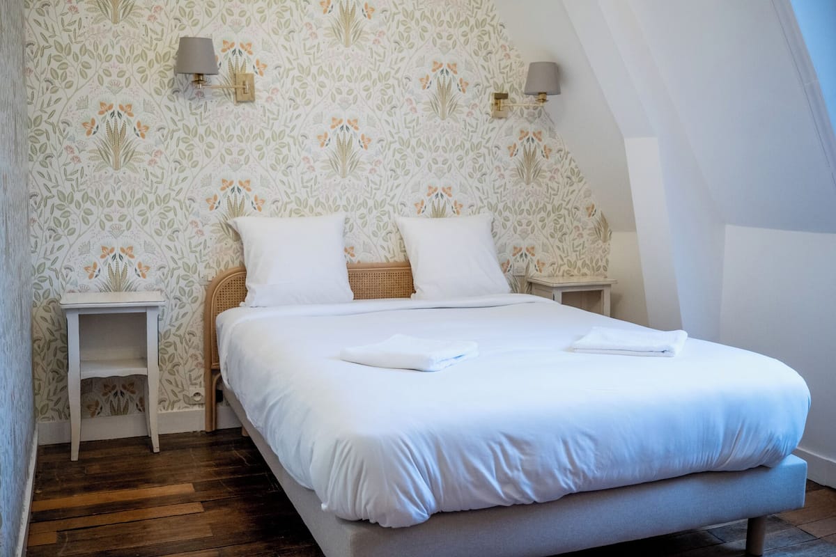 Een net opgemaakt bed in een kamer met bloemen behang, twee nachtkastjes met lampen, en een houten vloer.