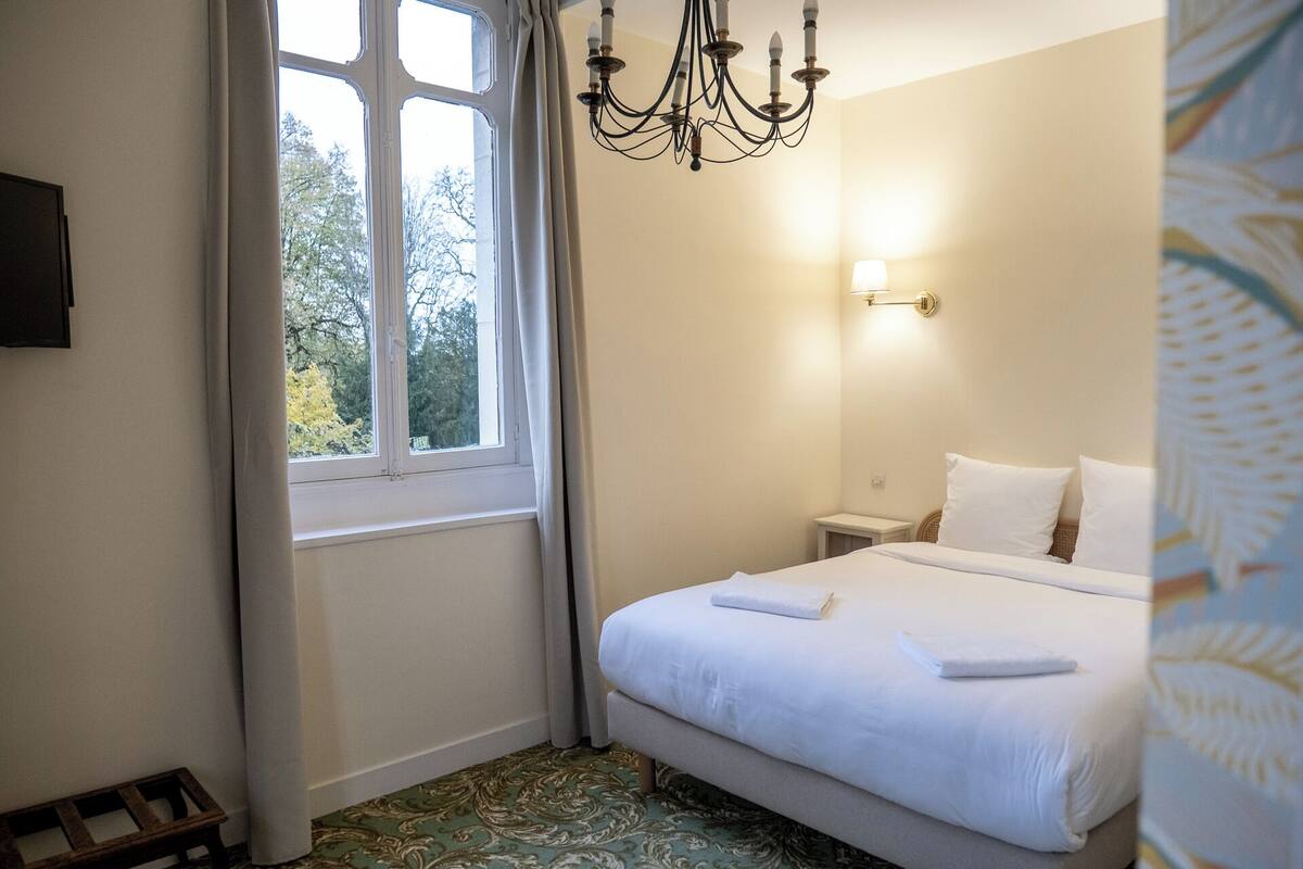 Een hotelkamer met een bed, nachtkastje, een wandlamp en een raam met gordijnen.