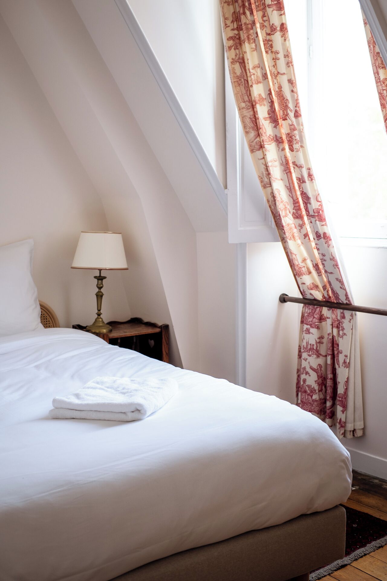 A neatly made bed in a room with a sloped ceiling, a bedside table with a lamp, and a window with floral curtains.