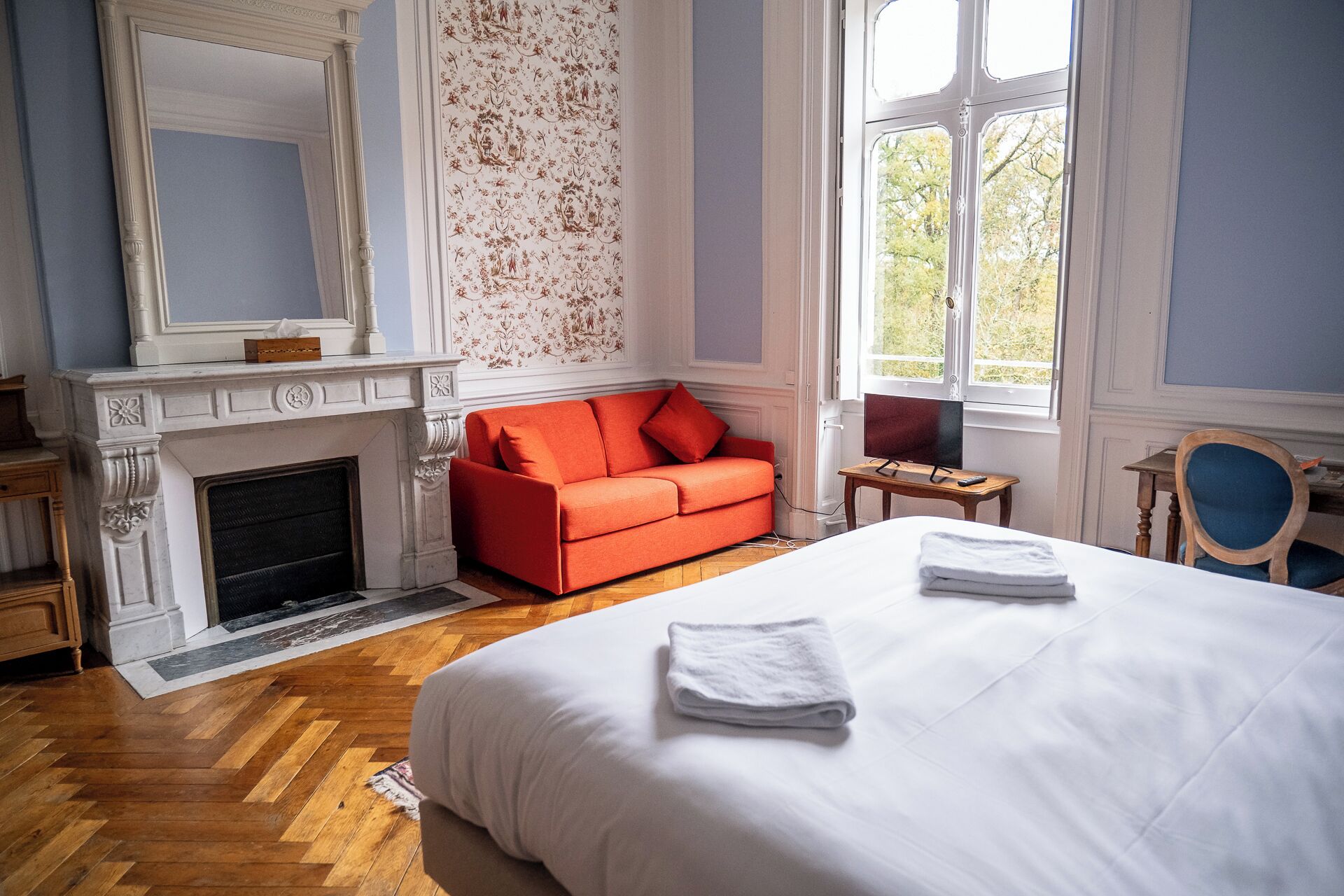A hotel room with a large bed, a fireplace, a red sofa, and a window with a view of trees.