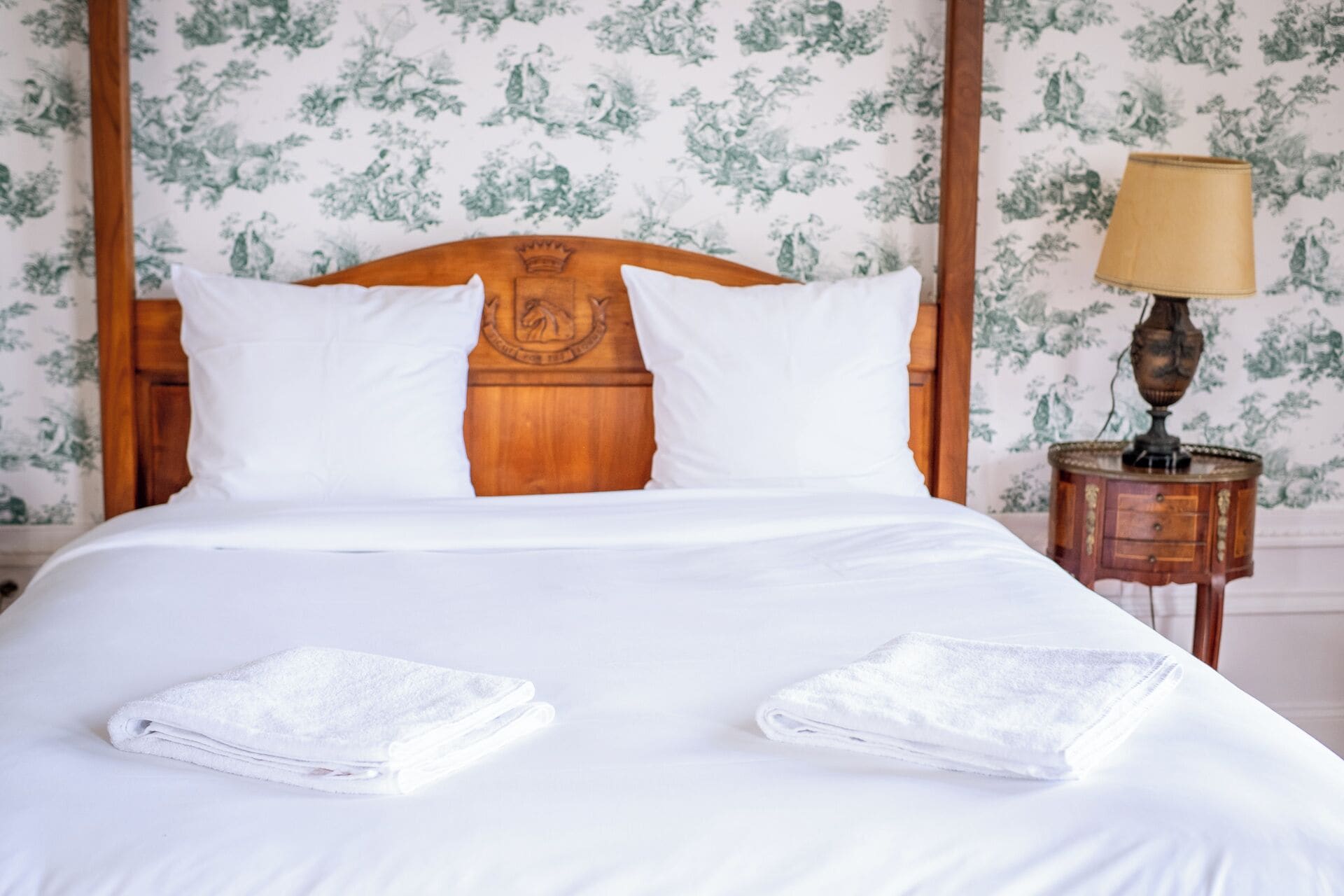 Un lit bien fait, avec du linge de lit blanc et des oreillers, une tête de lit en bois ornée d’un motif et une table de chevet avec une lampe et un petit tiroir.