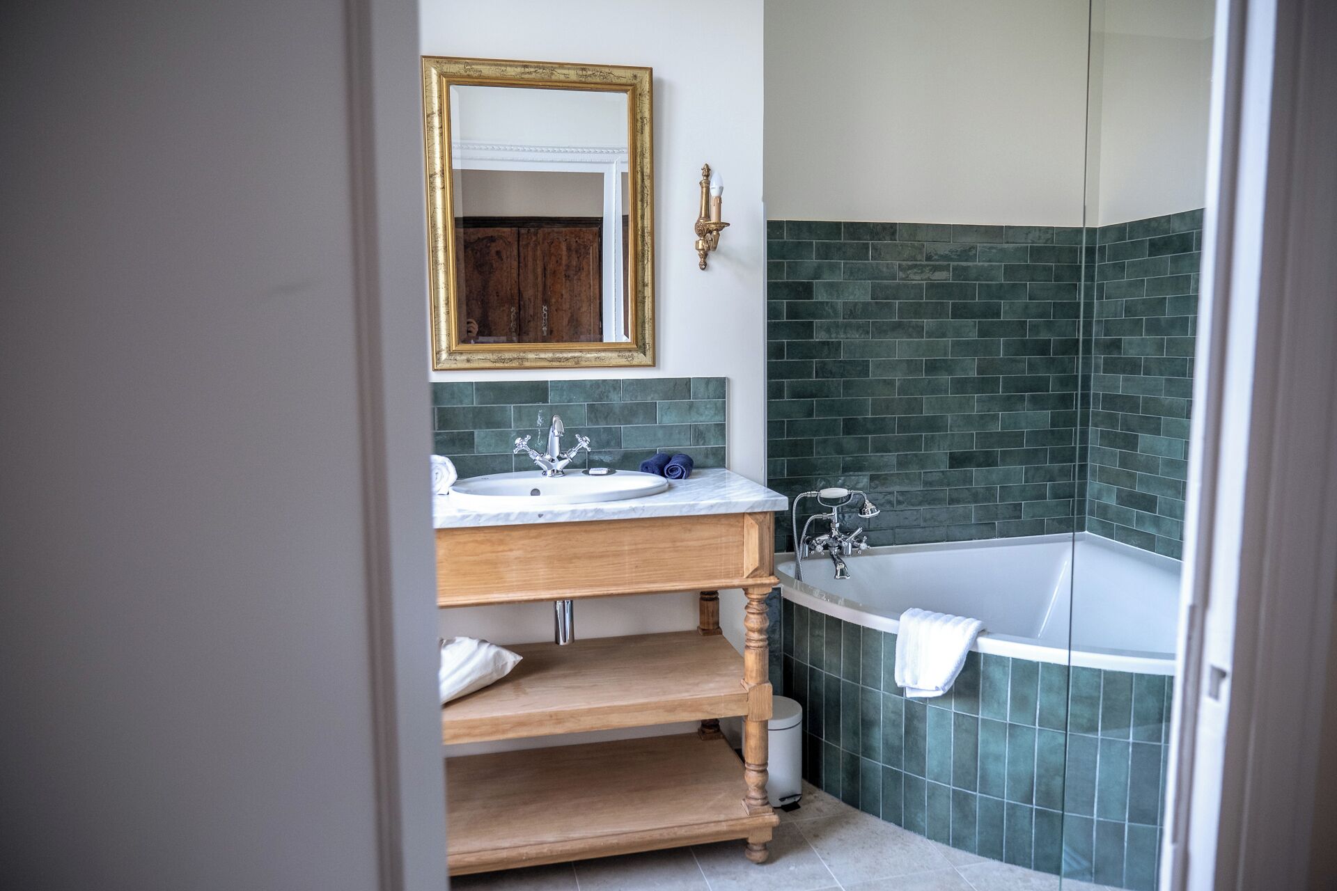A bathroom with a wooden vanity, a white sink, a mirror with a gold frame, and a green-tiled shower area.