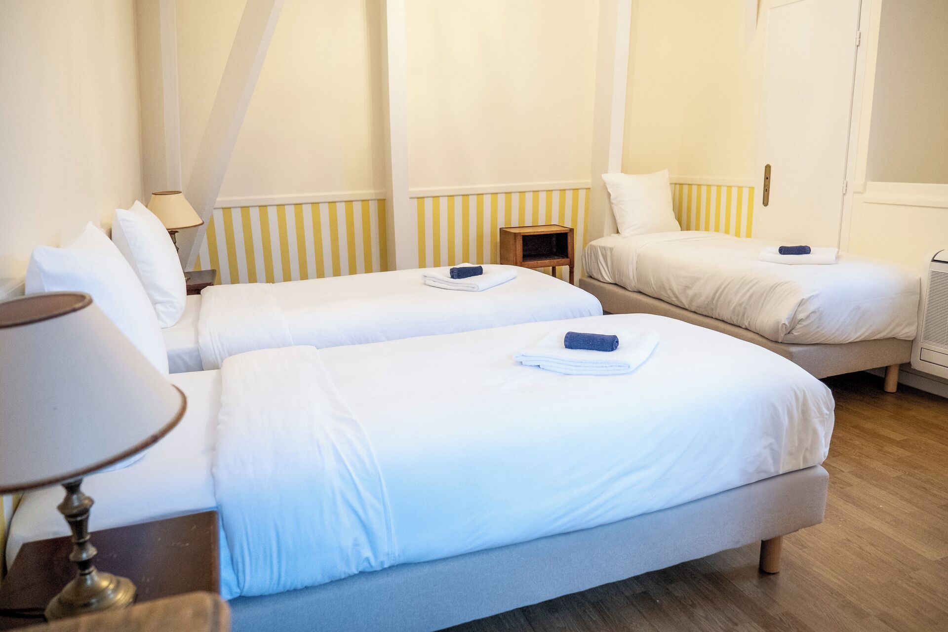 Habitación de hotel con dos camas individuales, una mesita de noche con lámpara, una pequeña mesita auxiliar de madera y un radiador.