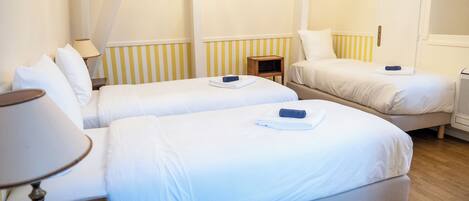 Habitación de hotel con dos camas individuales, una mesita de noche con lámpara, una pequeña mesita auxiliar de madera y un radiador.