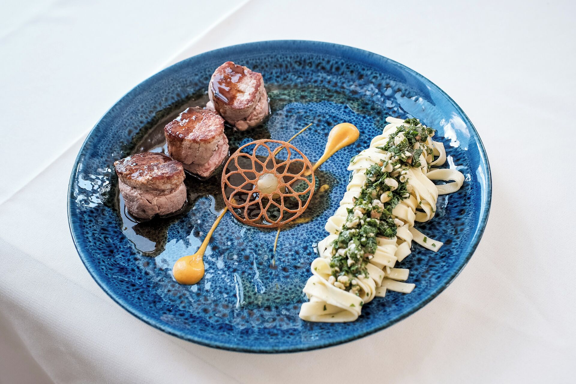A blue plate with three pieces of meat, a decorative item, and pasta garnished with herbs.