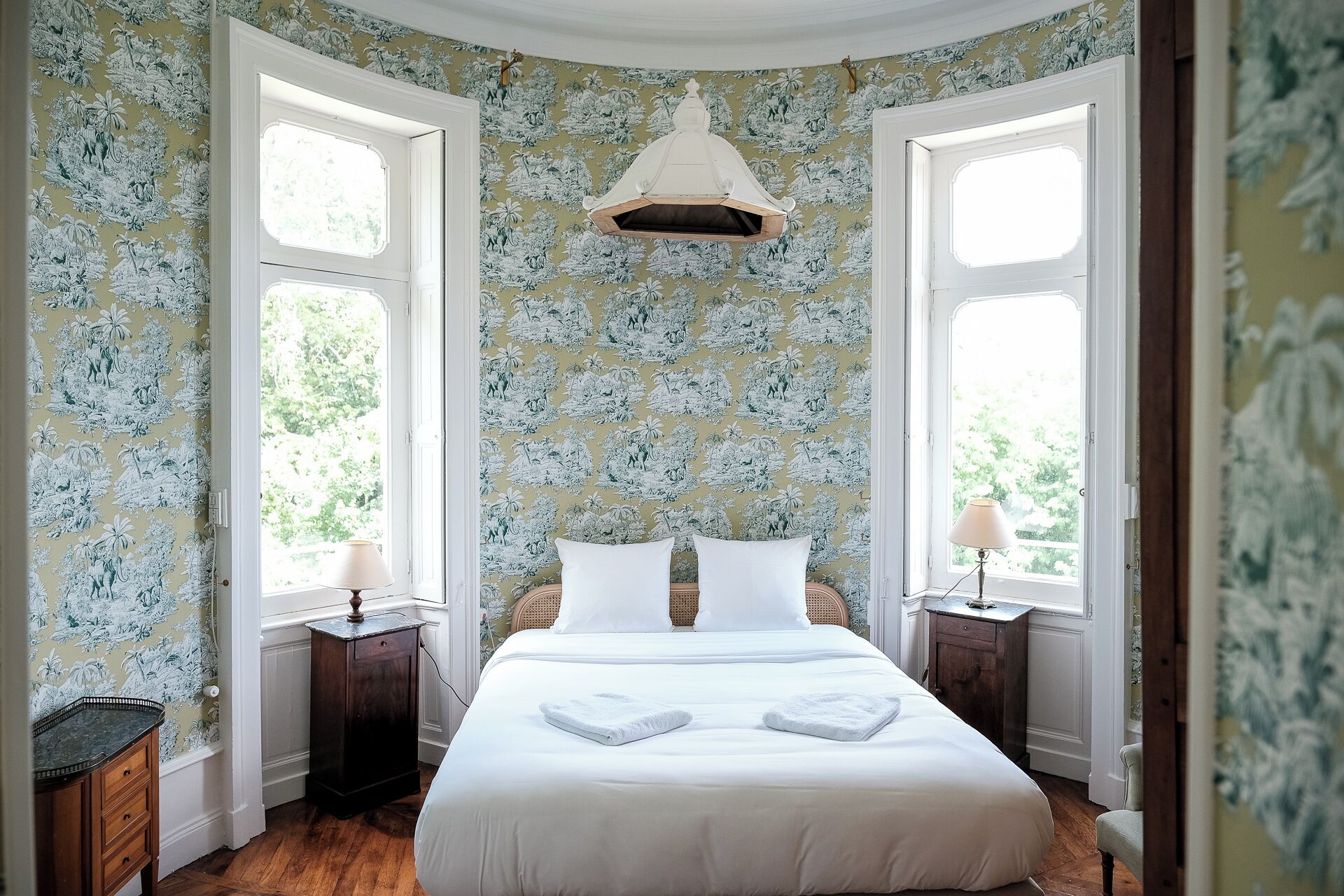 Ein Schlafzimmer mit einem großen Bett, zwei Nachttischen, einer Hängeleuchte und einem Fenster mit Blick auf Grünflächen.