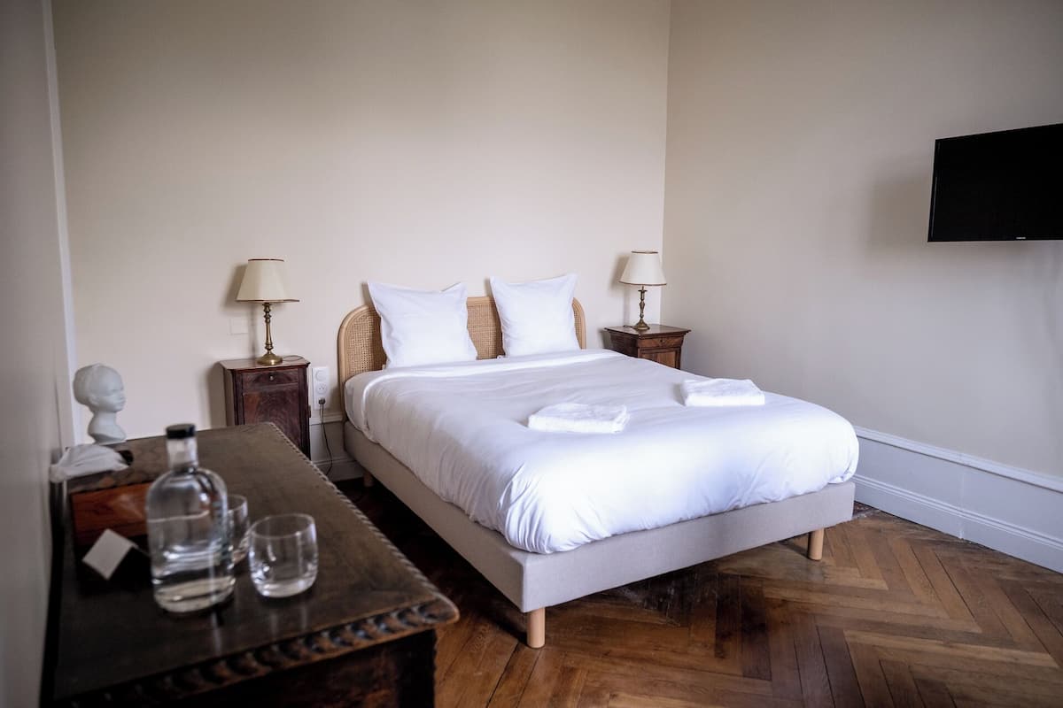 Een net opgemaakt bed met witte lakens, twee nachtkastjes met lampen, een houten nachtkastje, een tafel met marmeren blad en glazen, en een aan de muur bevestigde televisie.