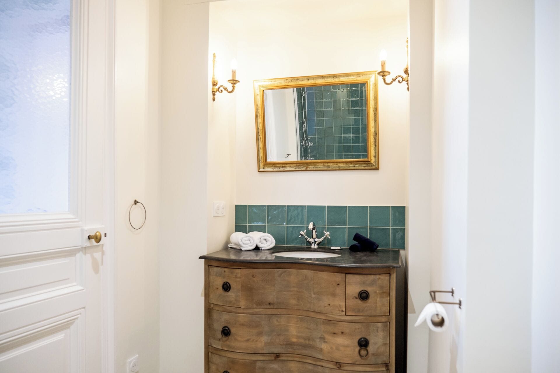 A bathroom with a wooden vanity, a mirror with a gold frame, and a green tiled backsplash.