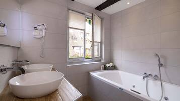 Un baño moderno con una bañera grande, dos lavamanos, un secador de pelo y una ventana con persianas.