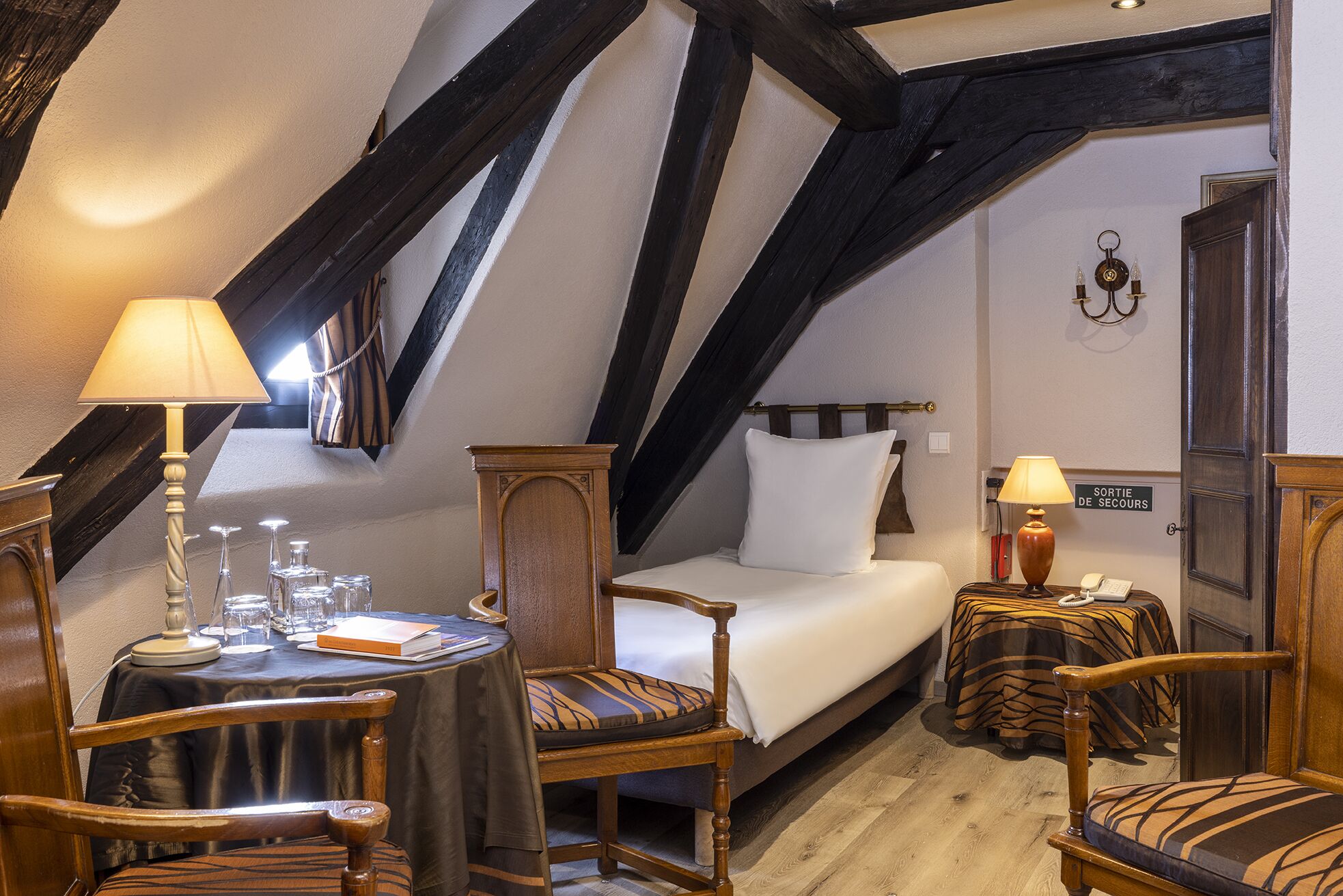 A cozy room with a bed, a table with a lamp, a chair, and a small table with a lamp. There is a sign on the wall that reads "SORTIE DE SECOURS".