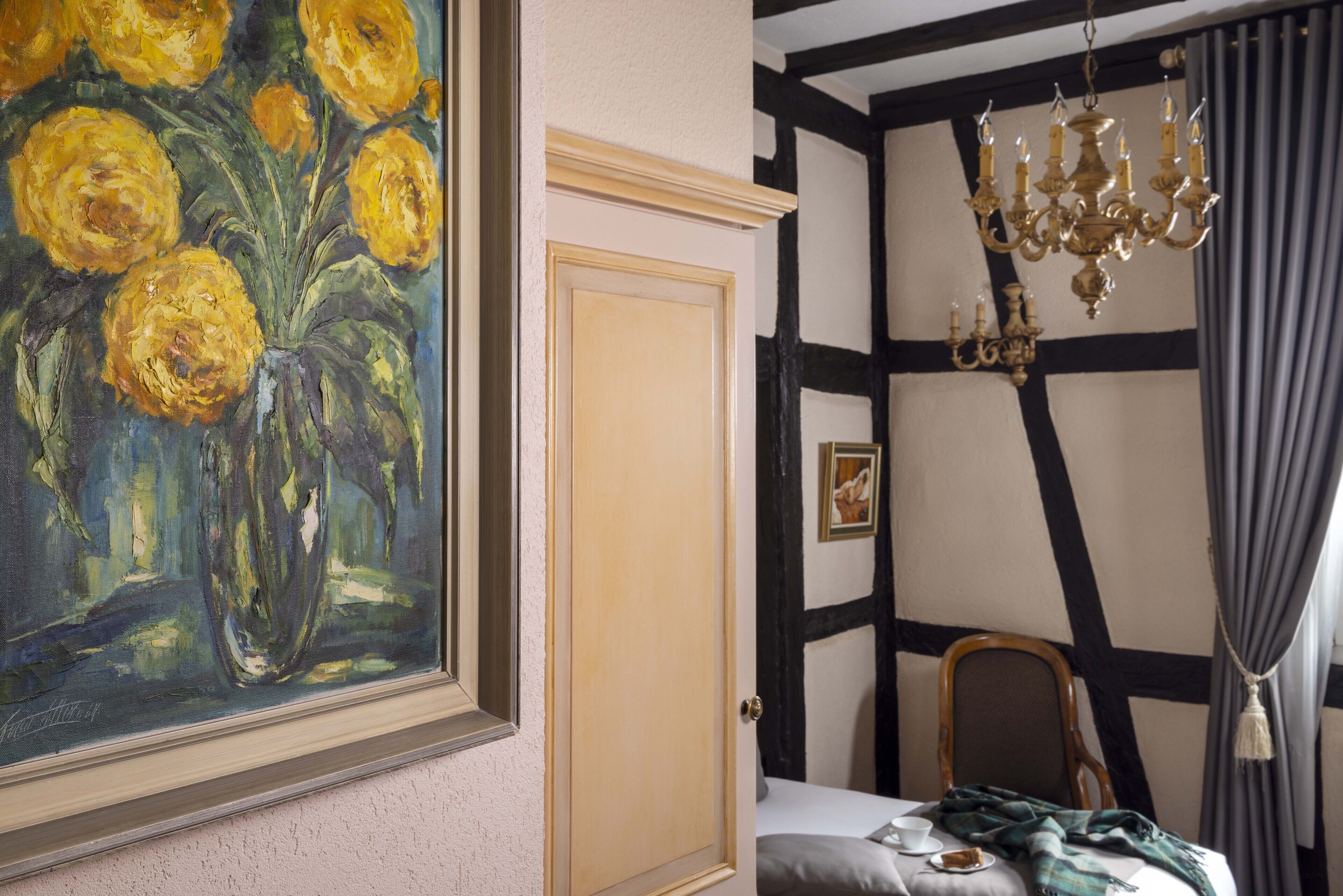 A room with a large painting of yellow flowers, a wooden door, a chandelier, and a bed with a blanket.
