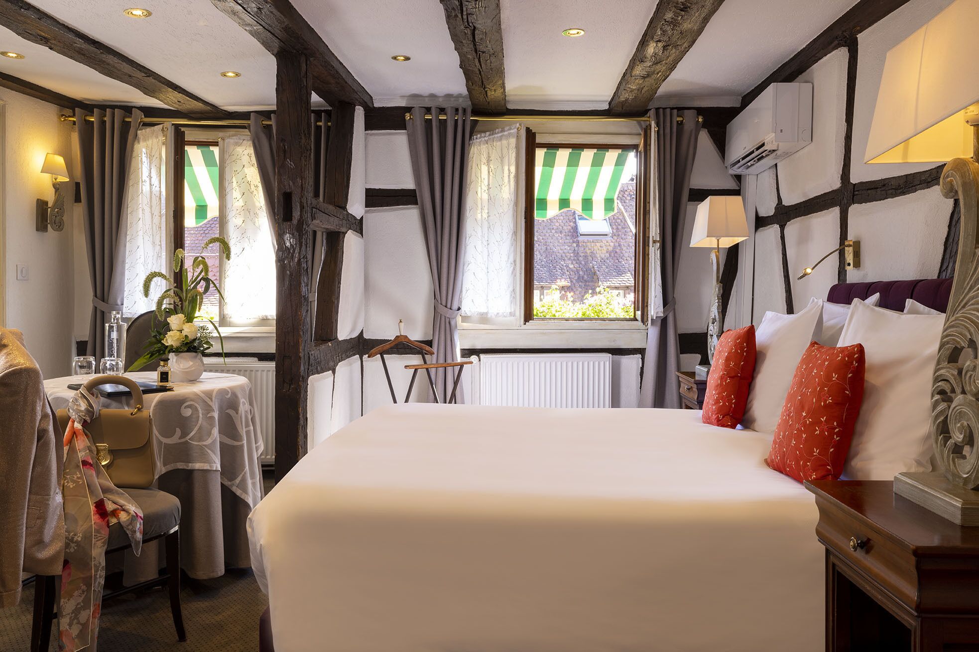 Une chambre d’hôtel avec un grand lit, une petite table avec une lampe, une chaise et une fenêtre avec des rideaux.