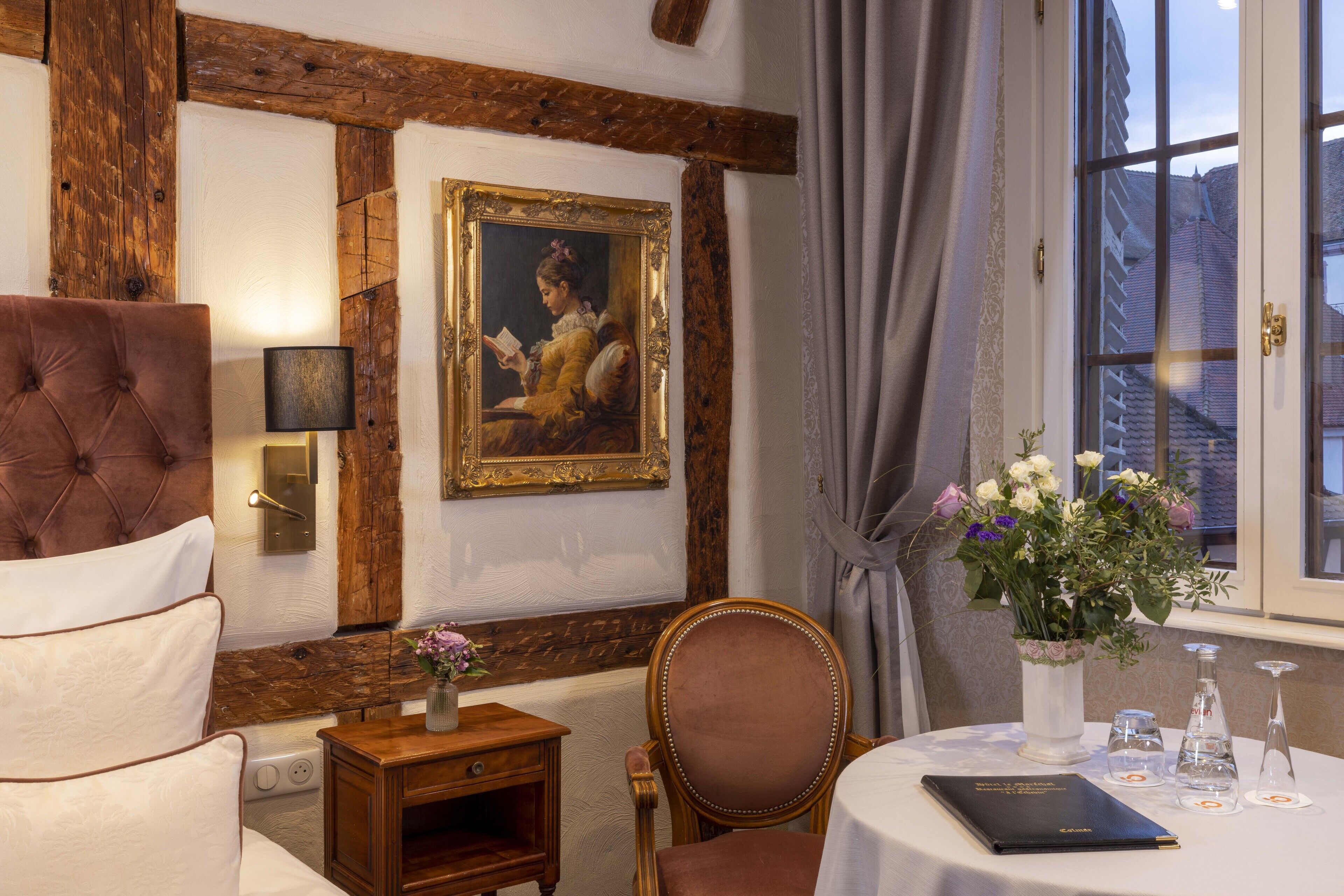 Una habitación con techo de madera, un cuadro enmarcado, una cama con cabecera, una mesa con un libro, un jarrón con flores y una ventana con cortinas.