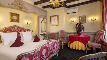 Ein Hotelzimmer mit einem großen Bett, zwei Sesseln, einem kleinen Tisch mit roter Tischdecke und einer Kristallleuchter.