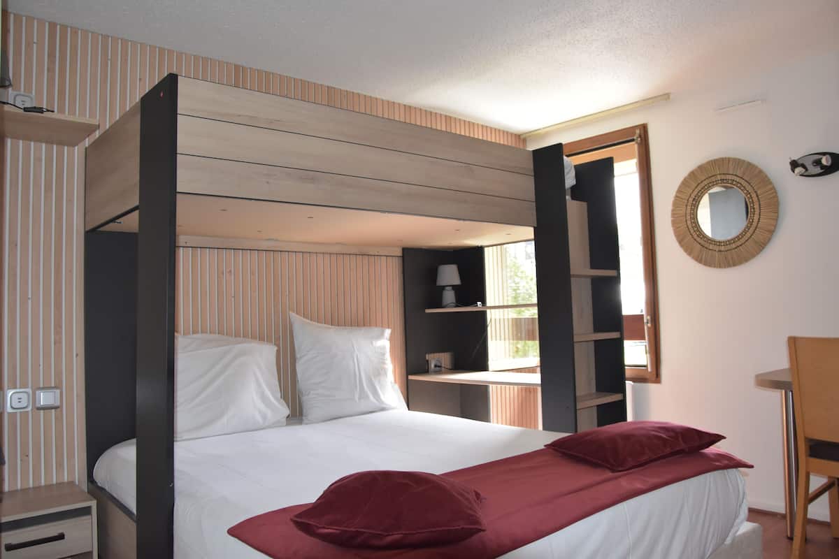 A bedroom with a bunk bed, a bedside table, a chair, a round mirror, and a window with blinds.