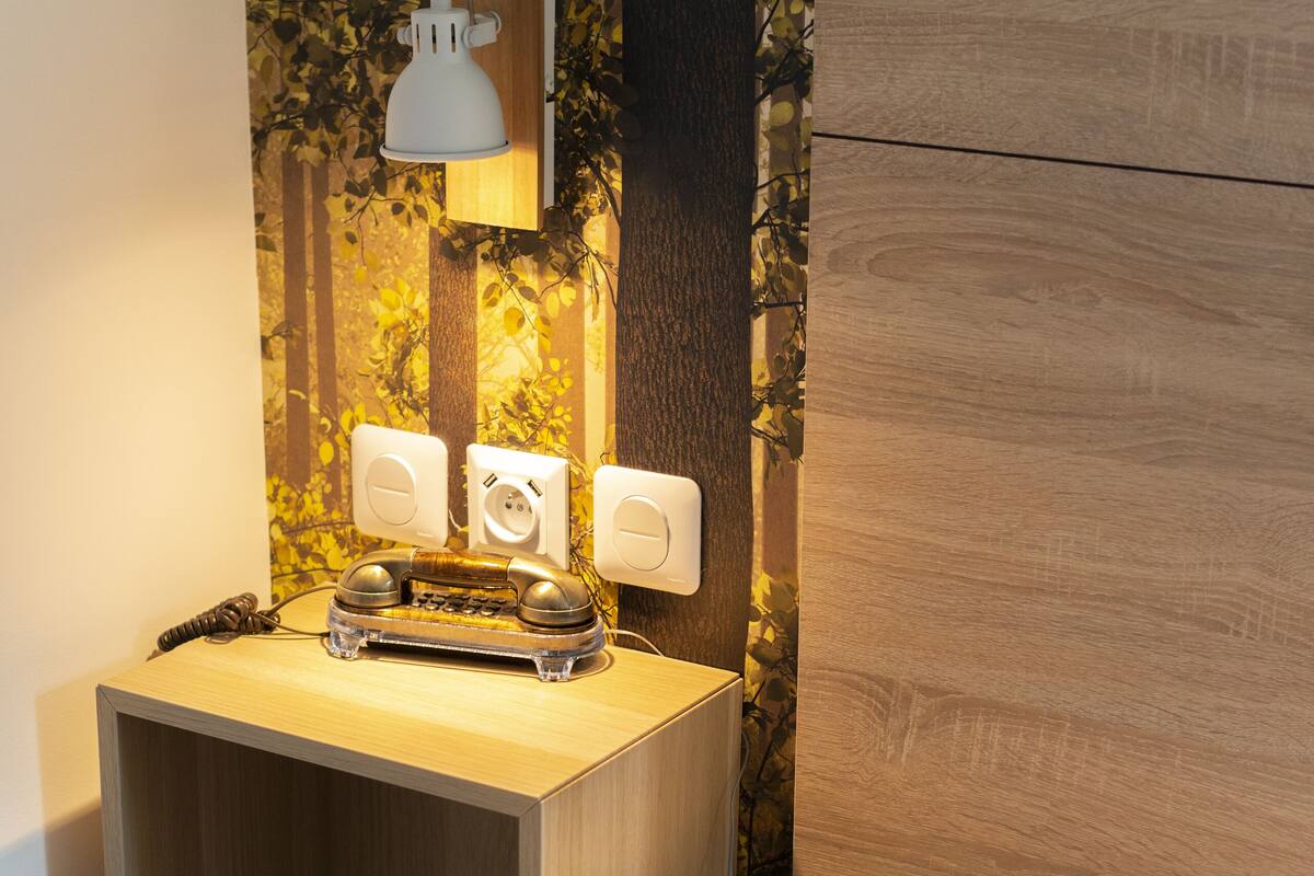 A neatly made bed with a wooden nightstand beside it, a vintage rotary phone, and a wall-mounted lamp.