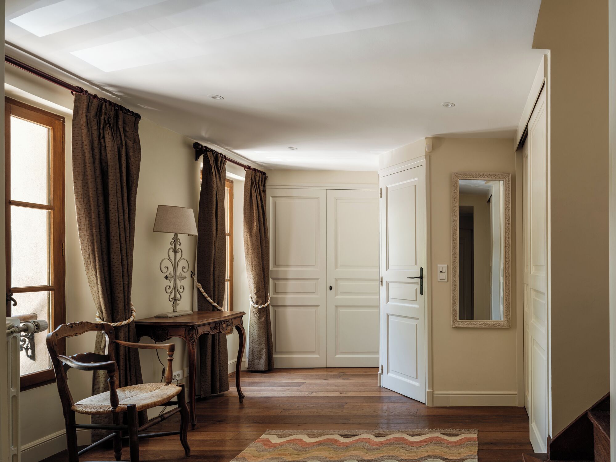 A room with a wooden floor, a chair, a table with a lamp, a door, and a mirror on the wall.
