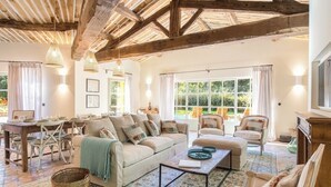 Amplia sala de estar con techo alto, vigas de madera, un conjunto de sofá, una mesa de comedor y vistas al jardín a través de grandes ventanales.