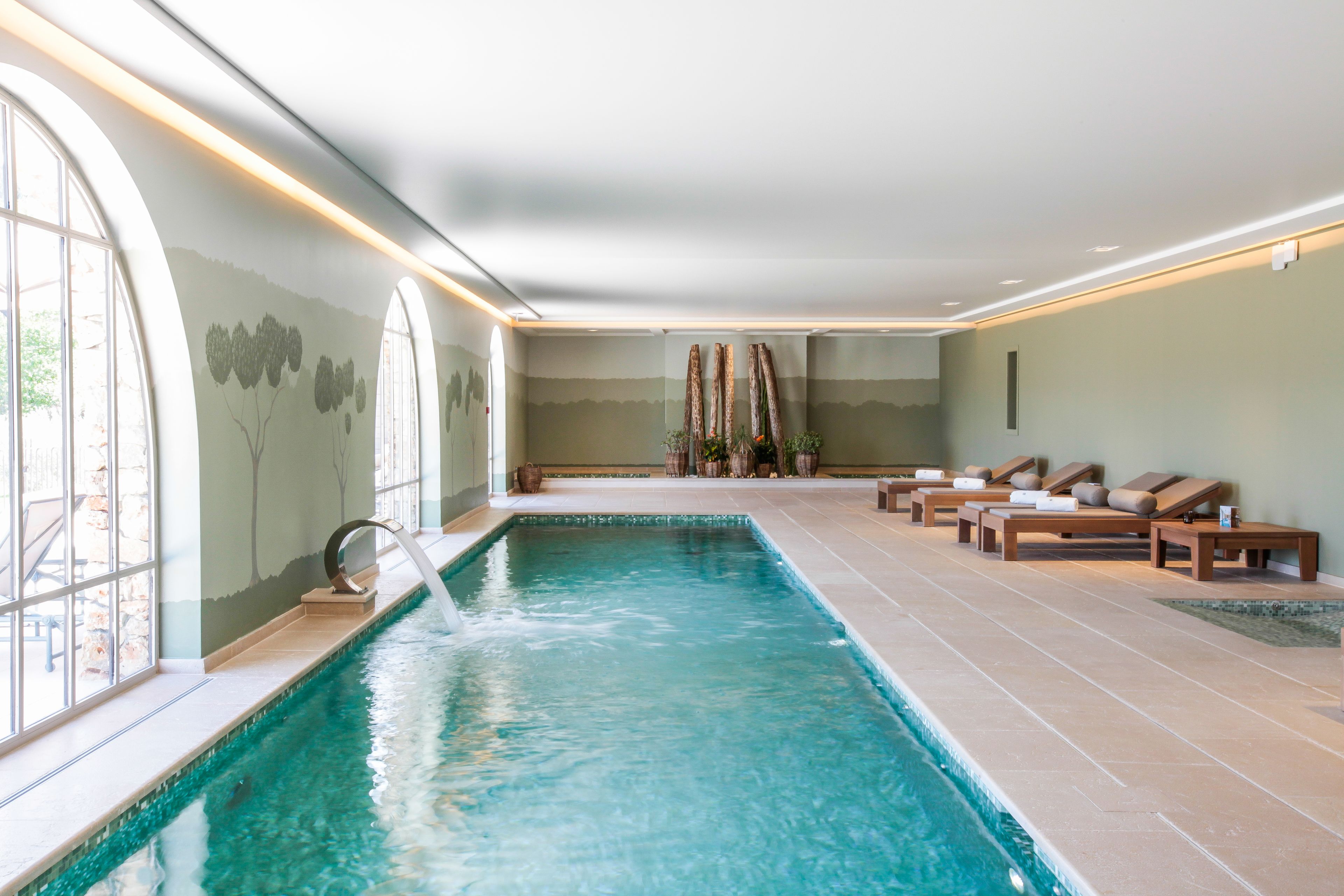 An indoor swimming pool with lounge chairs and a view of trees through large windows.