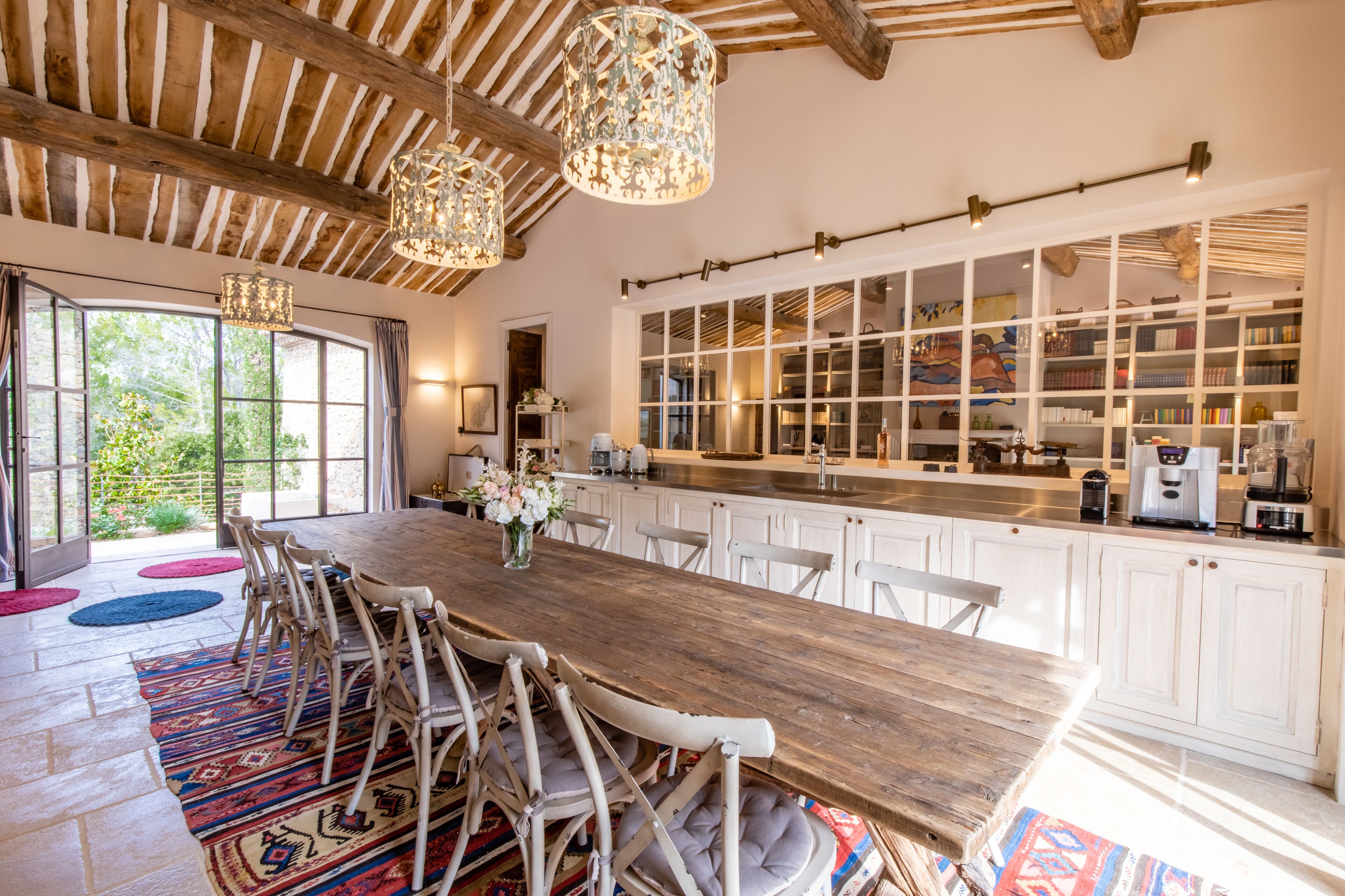 A spacious dining area with a wooden table and chairs, a large window with a view of greenery, and a kitchen with white cabinets and a built-in bookshelf.