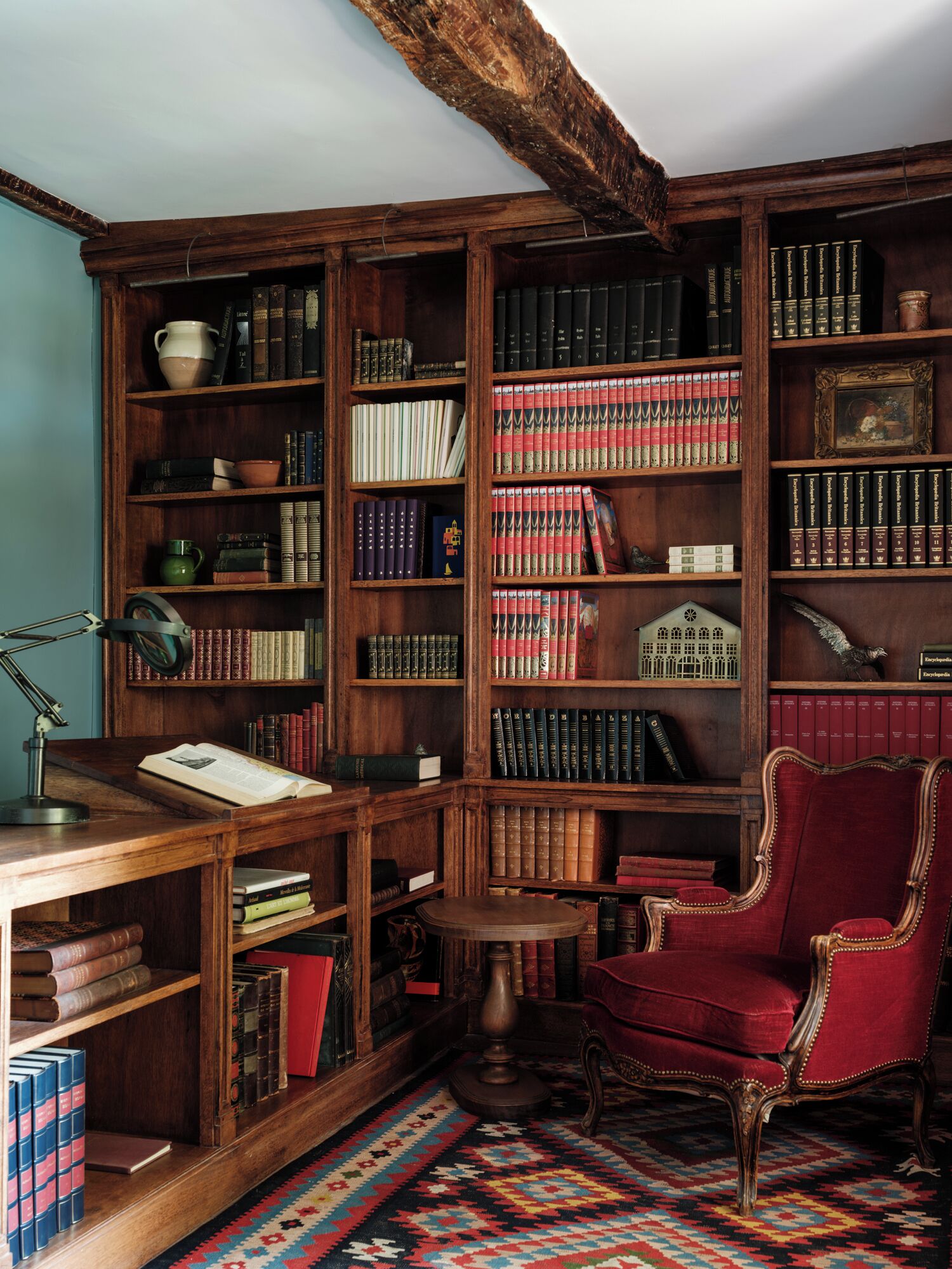 A library with wooden bookshelves, a desk with a lamp, a red chair, and a patterned rug.