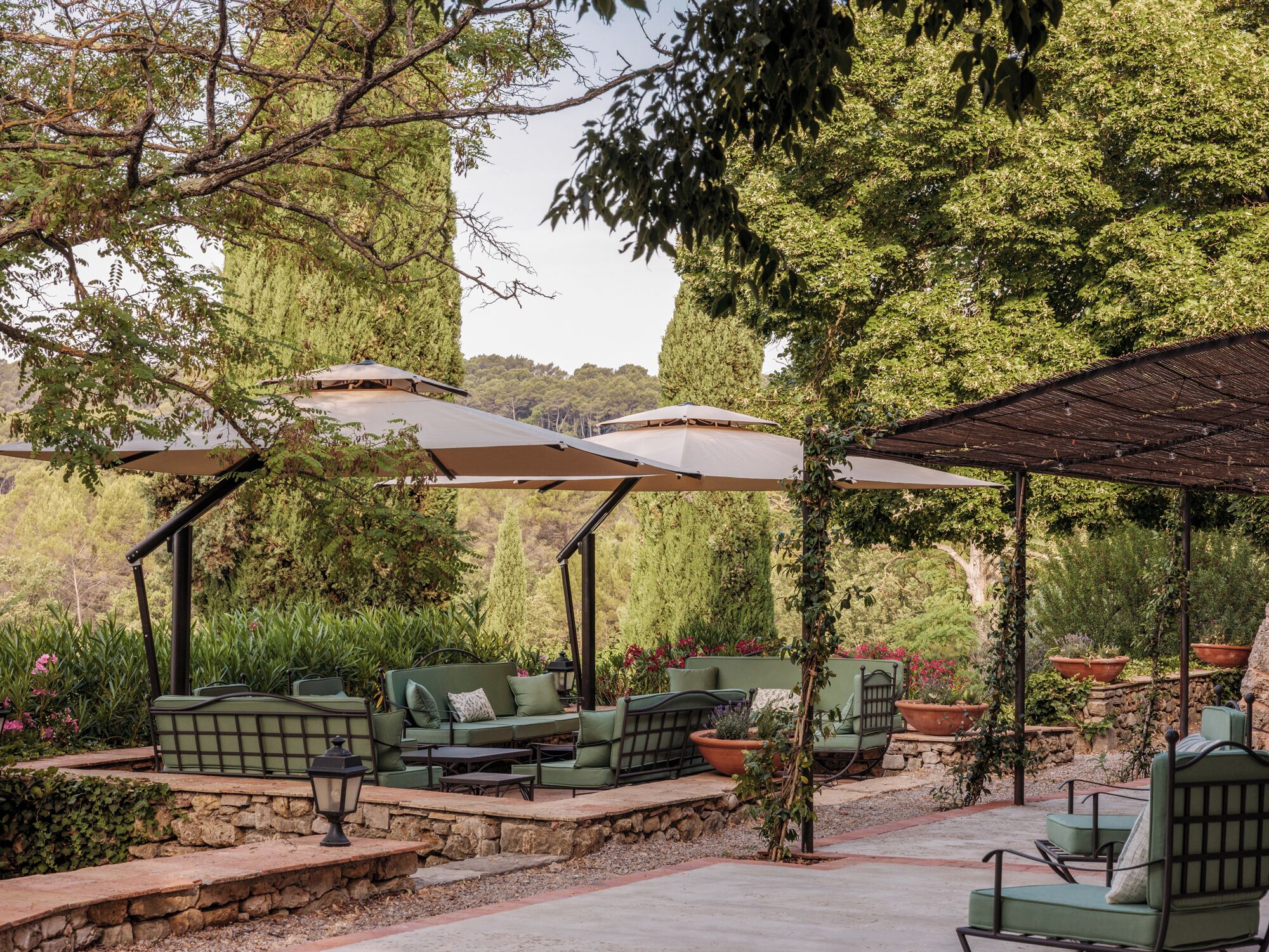 An outdoor seating area with green furniture and umbrellas, surrounded by greenery and stone walls.