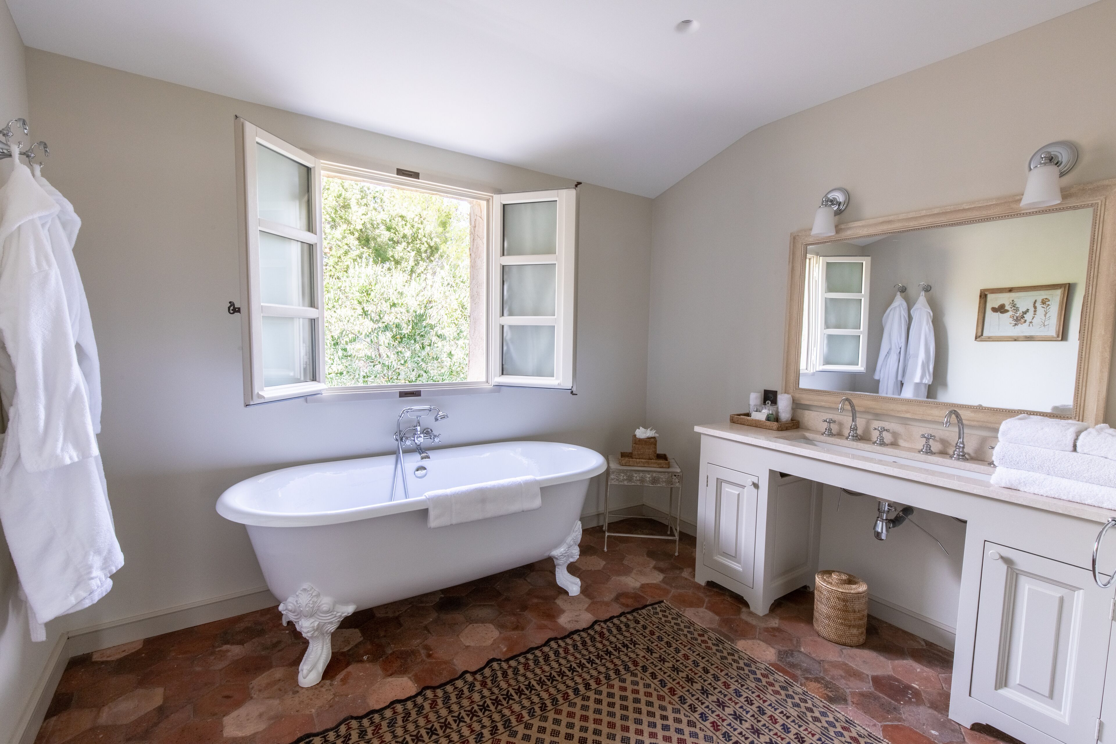 Un baño con una bañera independiente, un tocador con espejo y una ventana con contraventanas abiertas.